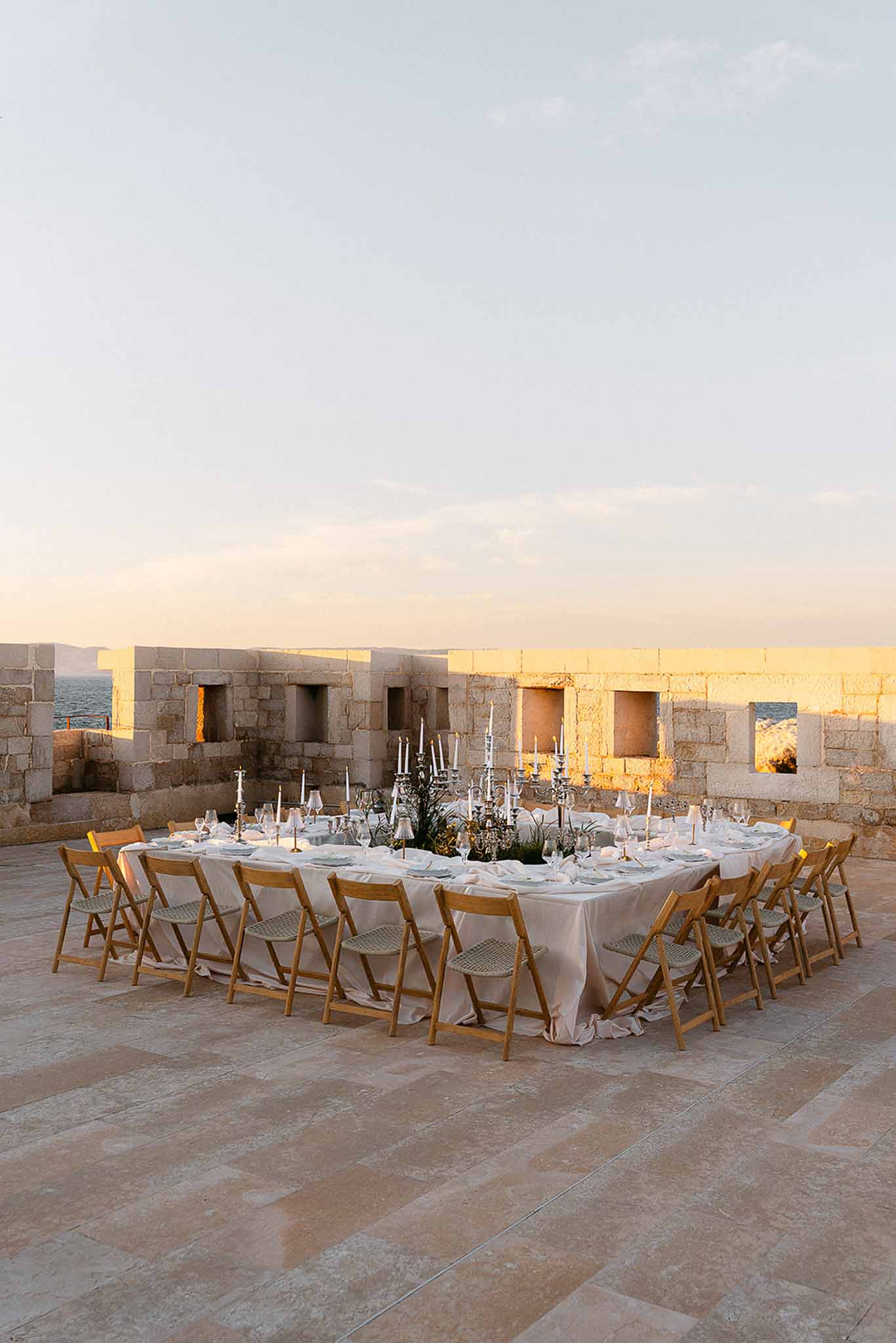 Outdoor reception table on stone terrace at sunset with silver candelabras, blush linen, and sea backdrop