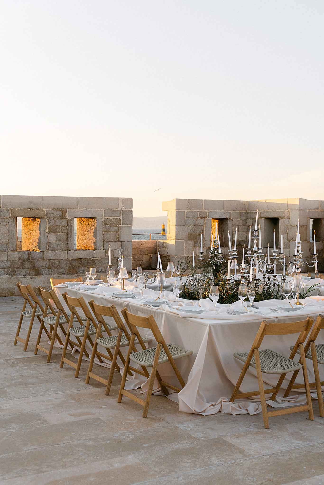 Long reception table on stone terrace with silver candelabras and coastal sunset view through crenellated walls