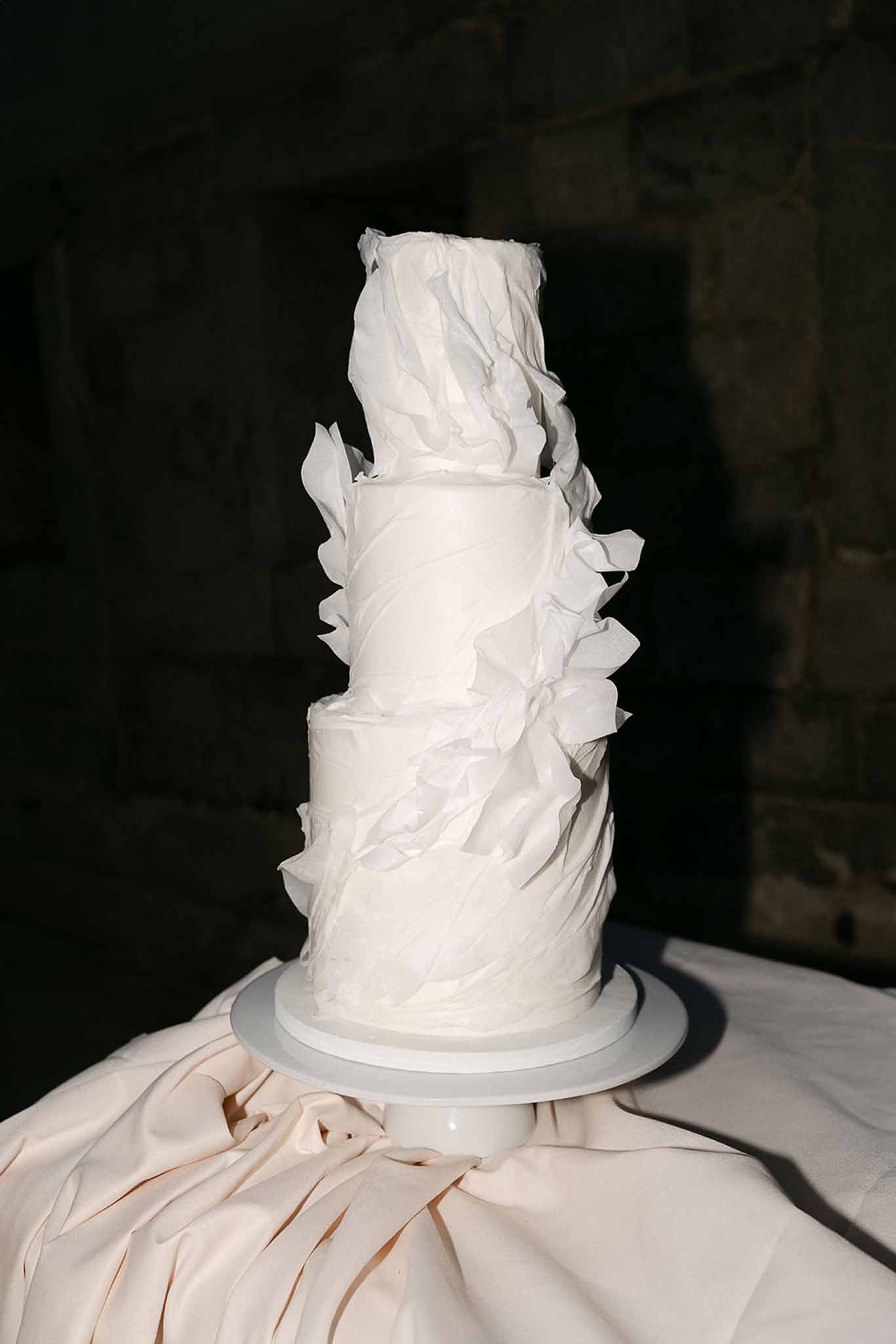 Three-tier white wedding cake with sculptural wafer paper ruffles on a pedestal in a stone-walled room
