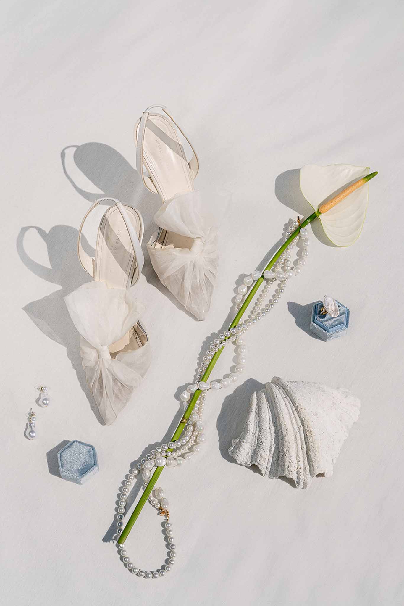 Bridal accessory flat lay with tulle bow heels, pearl necklace, blue velvet ring boxes, and pearl earrings