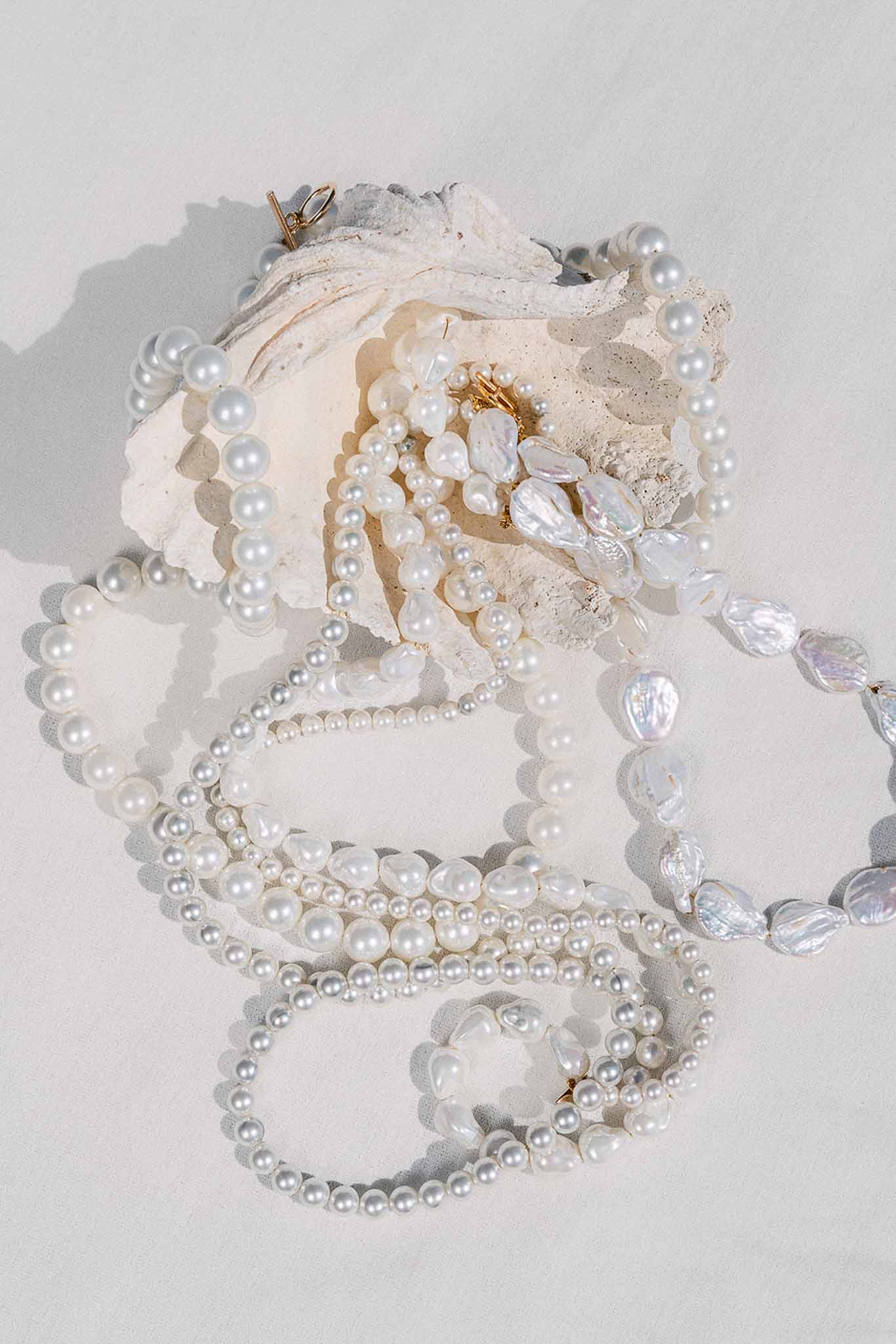 Pearl necklaces and bracelets arranged around white oyster shell on white surface