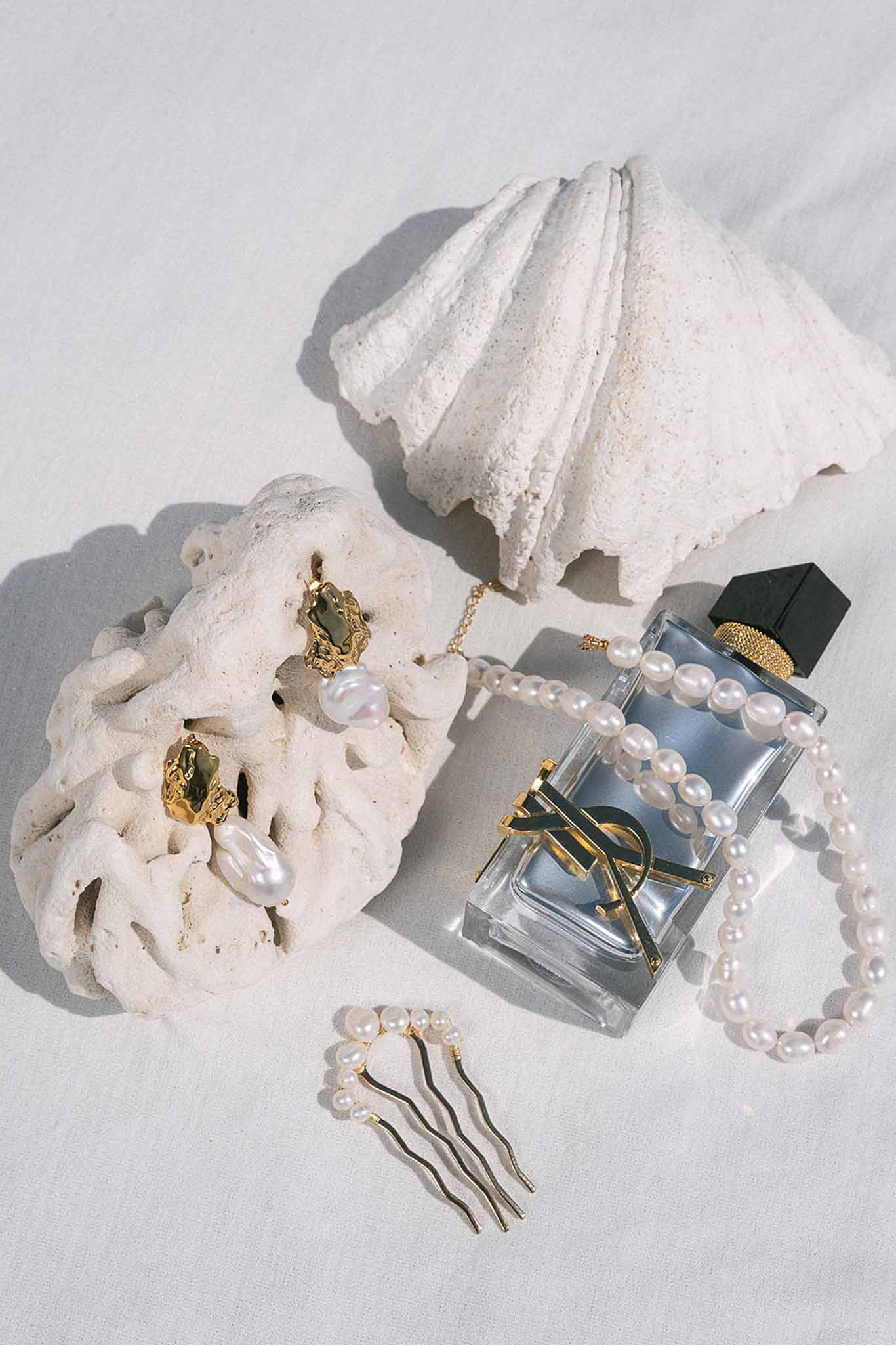 Flat lay of gold pearl earrings, pearl necklace, gold hair pins, and YSL perfume on white linen with coral props