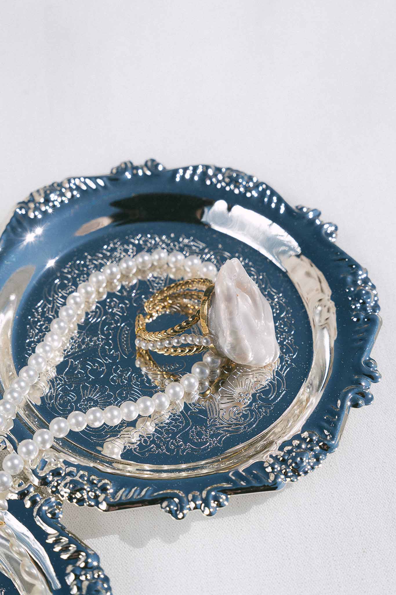Pearl necklace, gold twisted rings, and pink baroque stone ring on ornate navy enamel silver tray