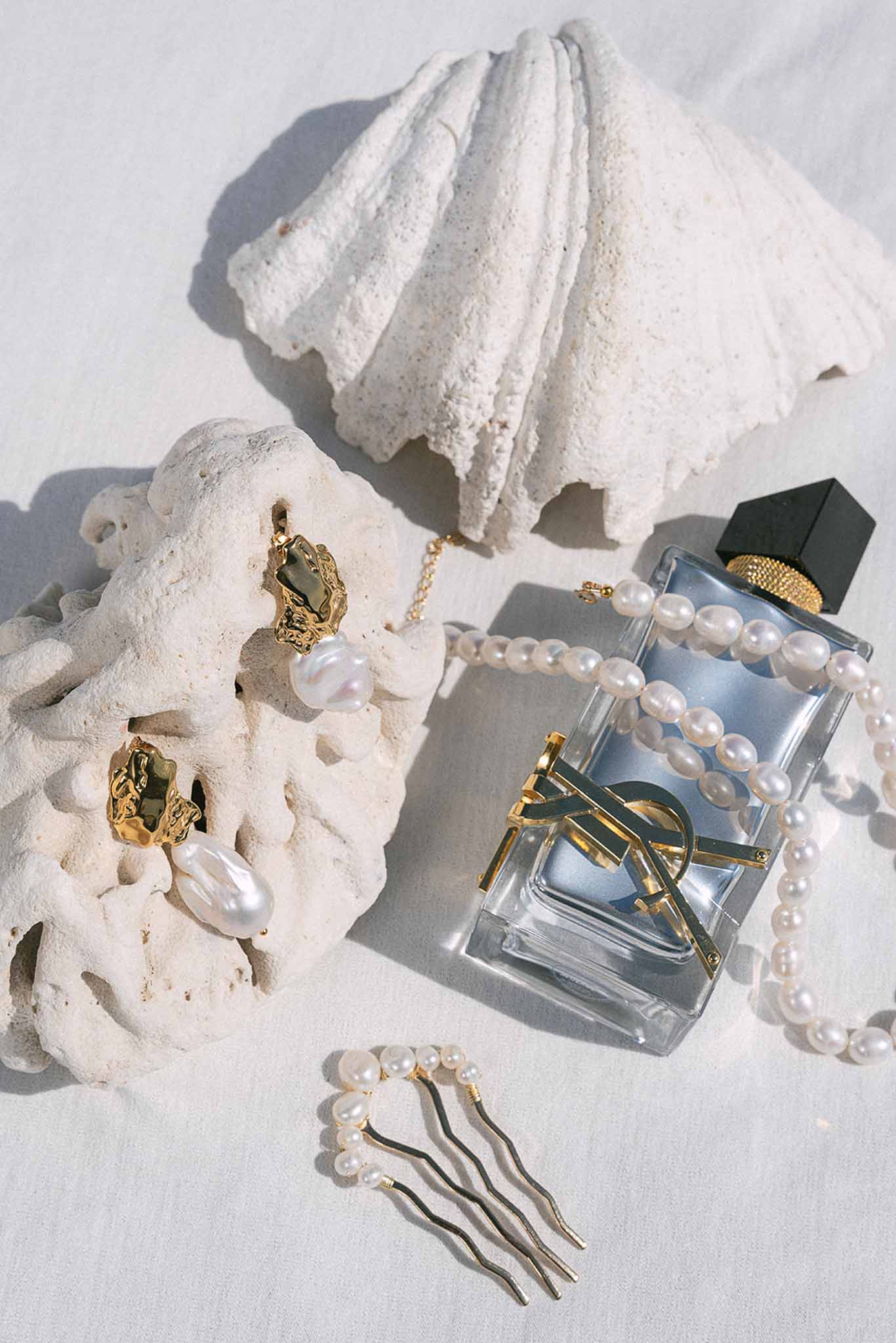 Pearl earrings, strand necklace, hair comb, and YSL perfume with white coral pieces on linen