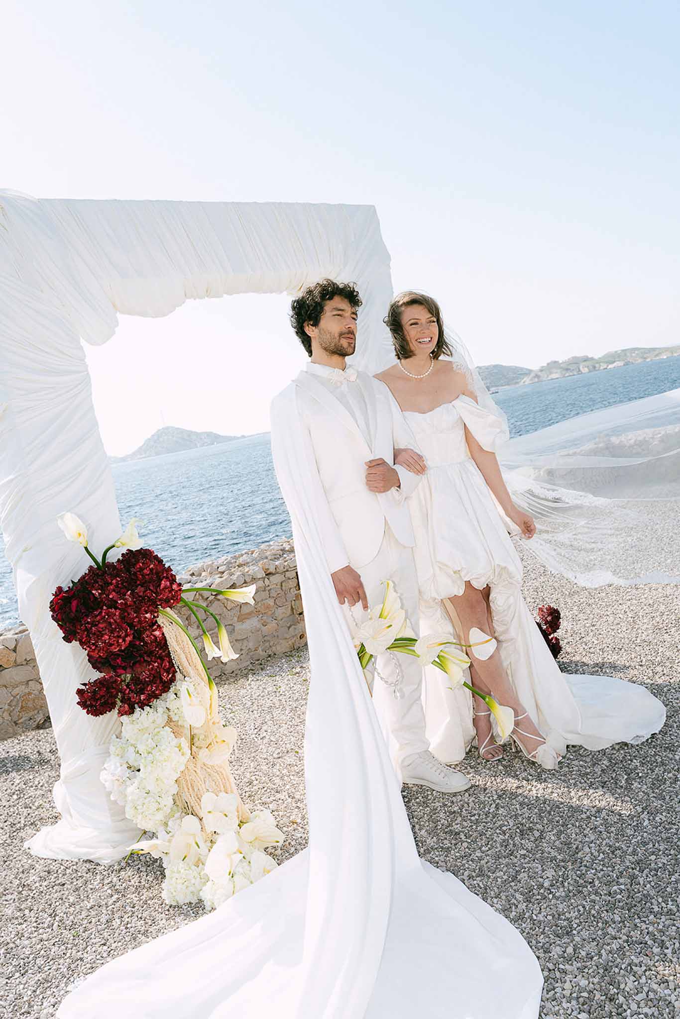 Couple under white fabric arch with burgundy hydrangea florals at coastal ceremony with ocean backdrop