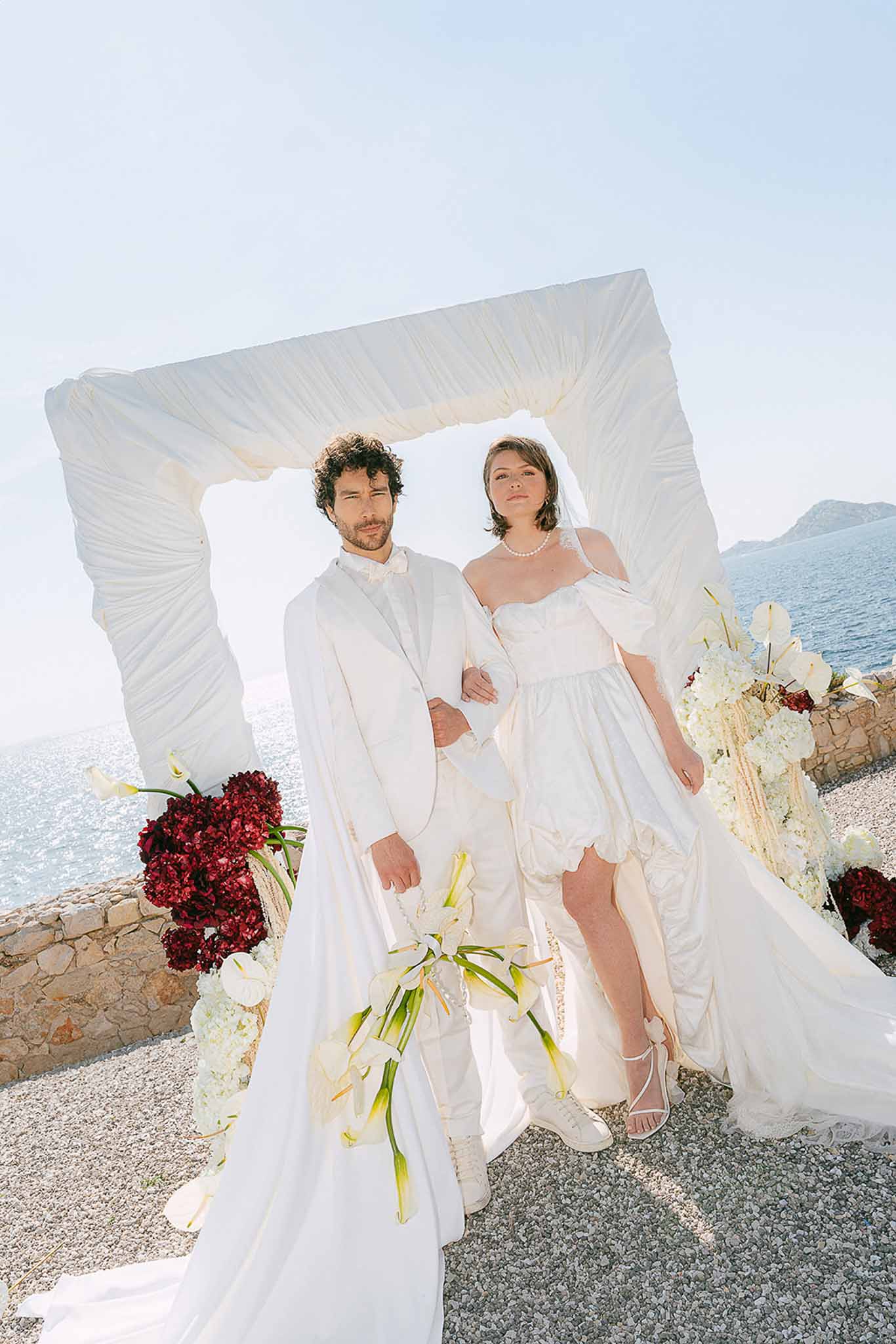 Couple posing under draped ivory arch with burgundy and white floral arrangements at coastal ceremony