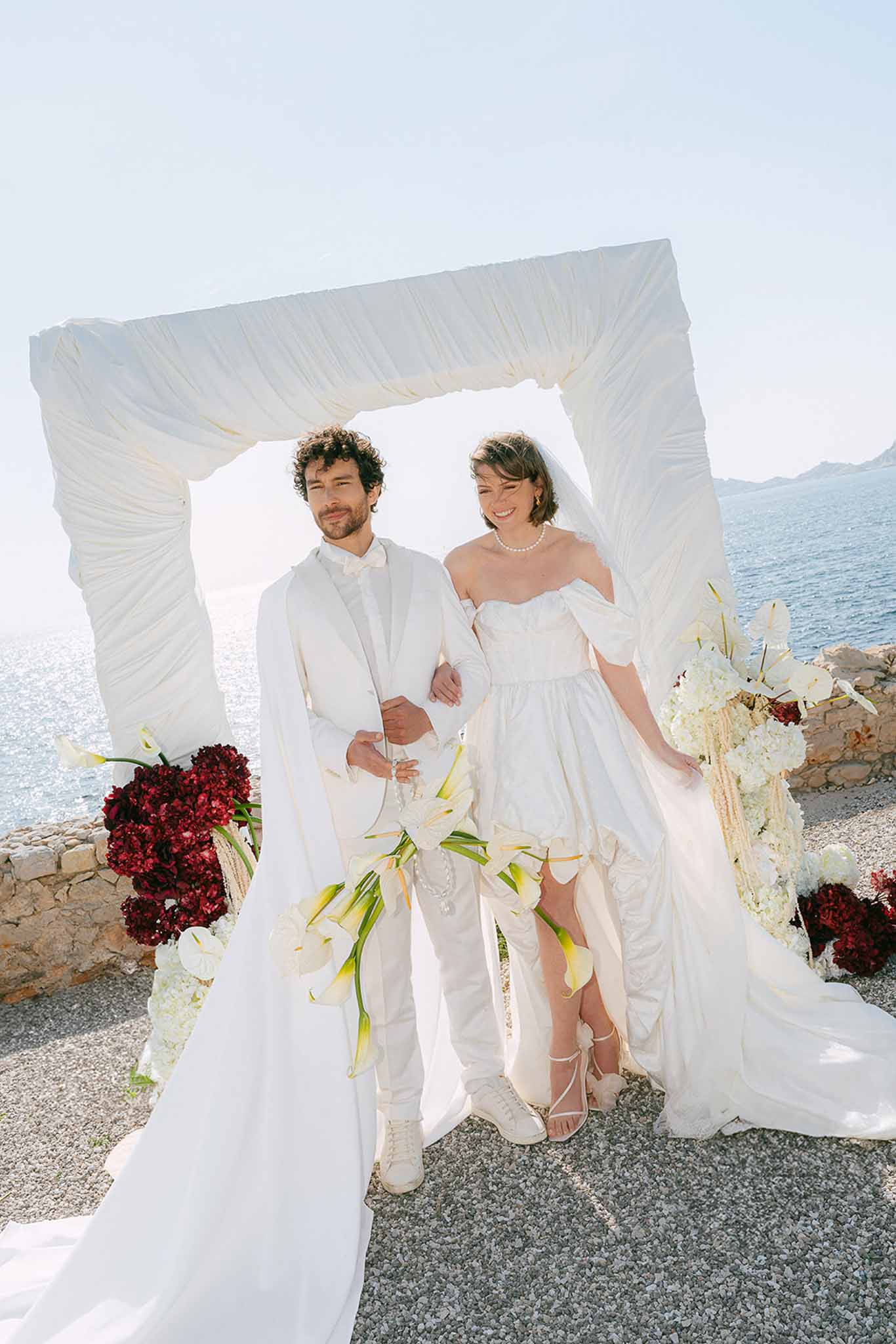 Couple under ivory-draped coastal arch with burgundy and white hydrangea and calla lily arrangements