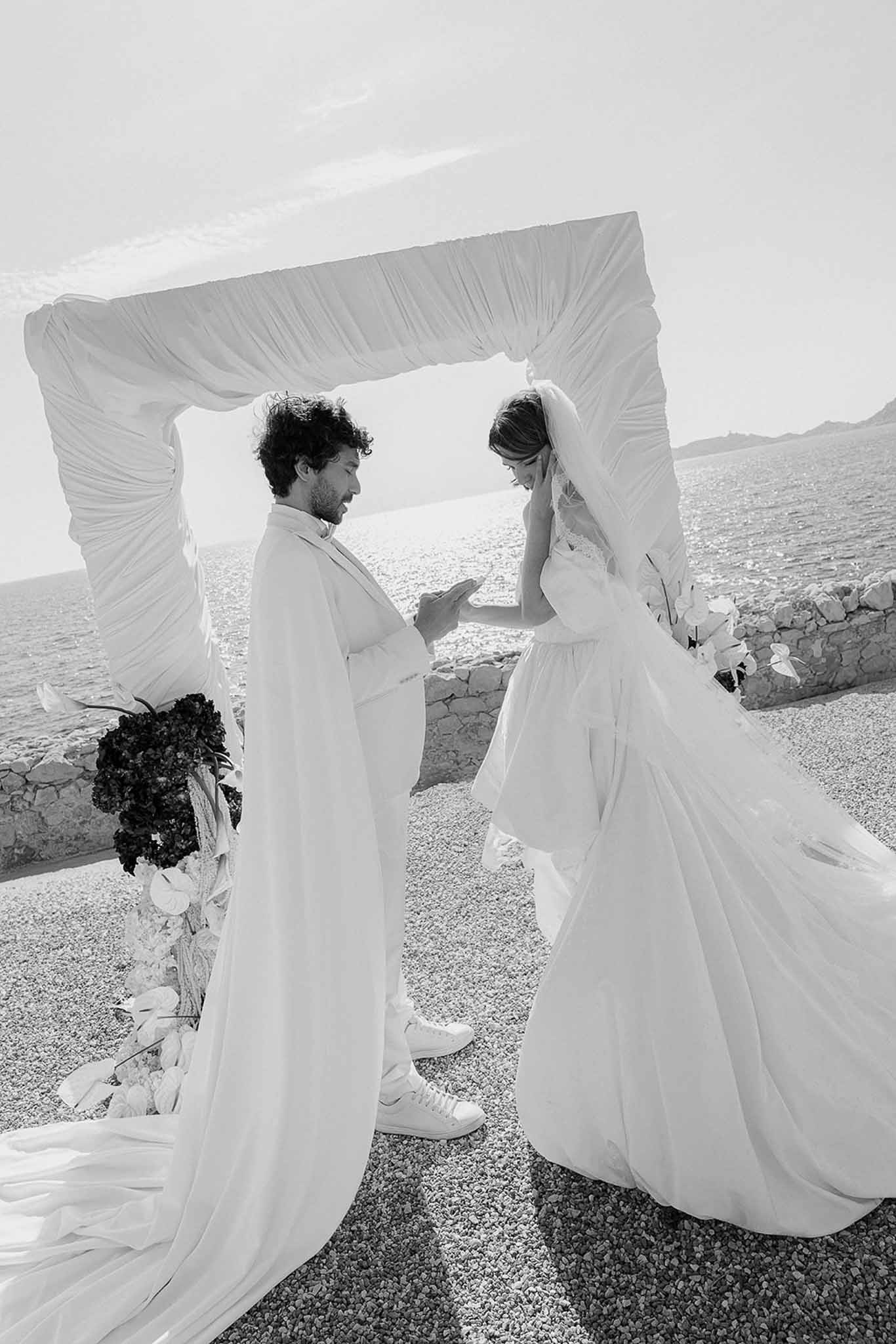 Black-and-white photo of couple exchanging rings under draped white fabric arch beside the sea