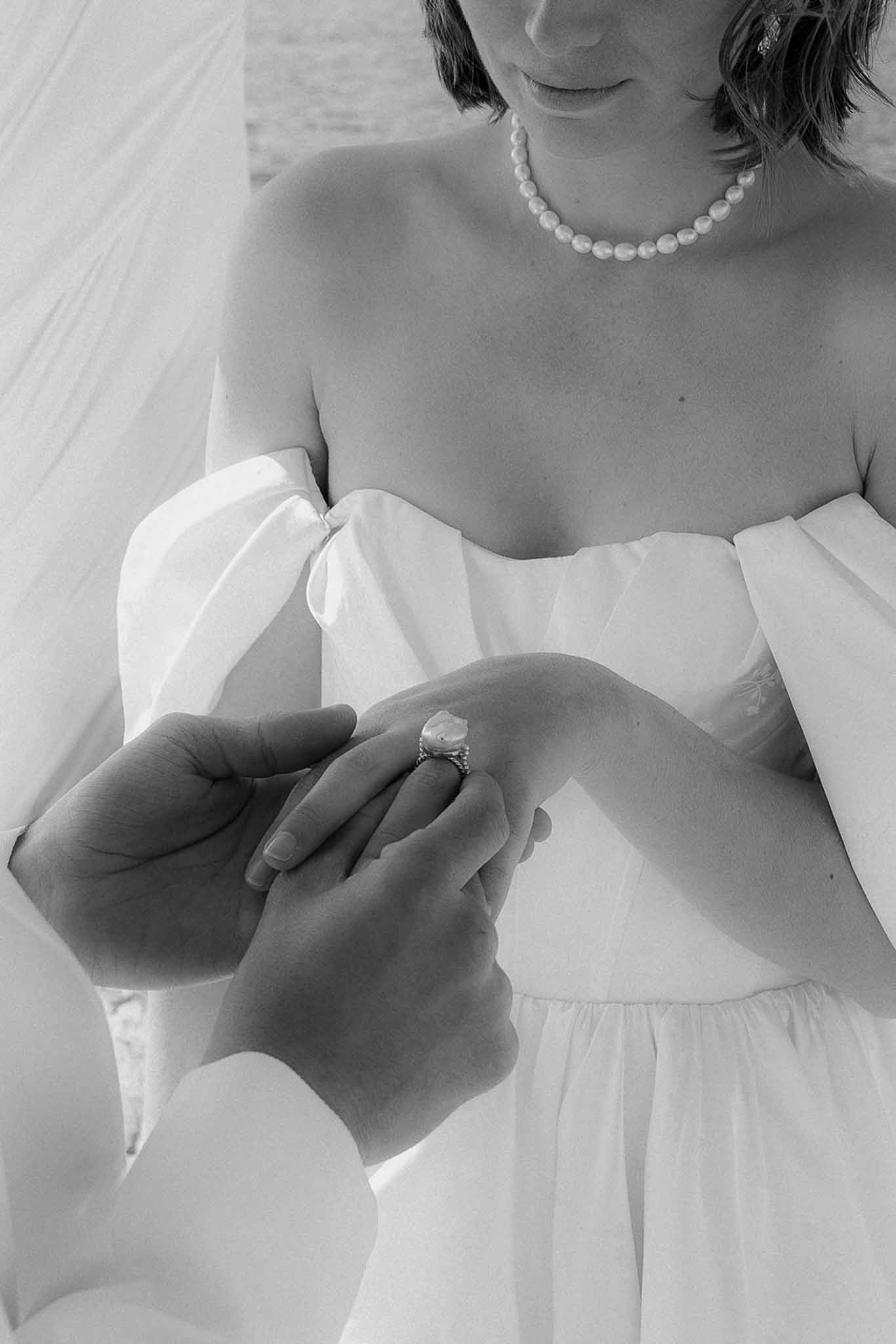 Black-and-white close-up of groom placing ring on bride's finger with pearl necklace and puff-sleeve gown visible