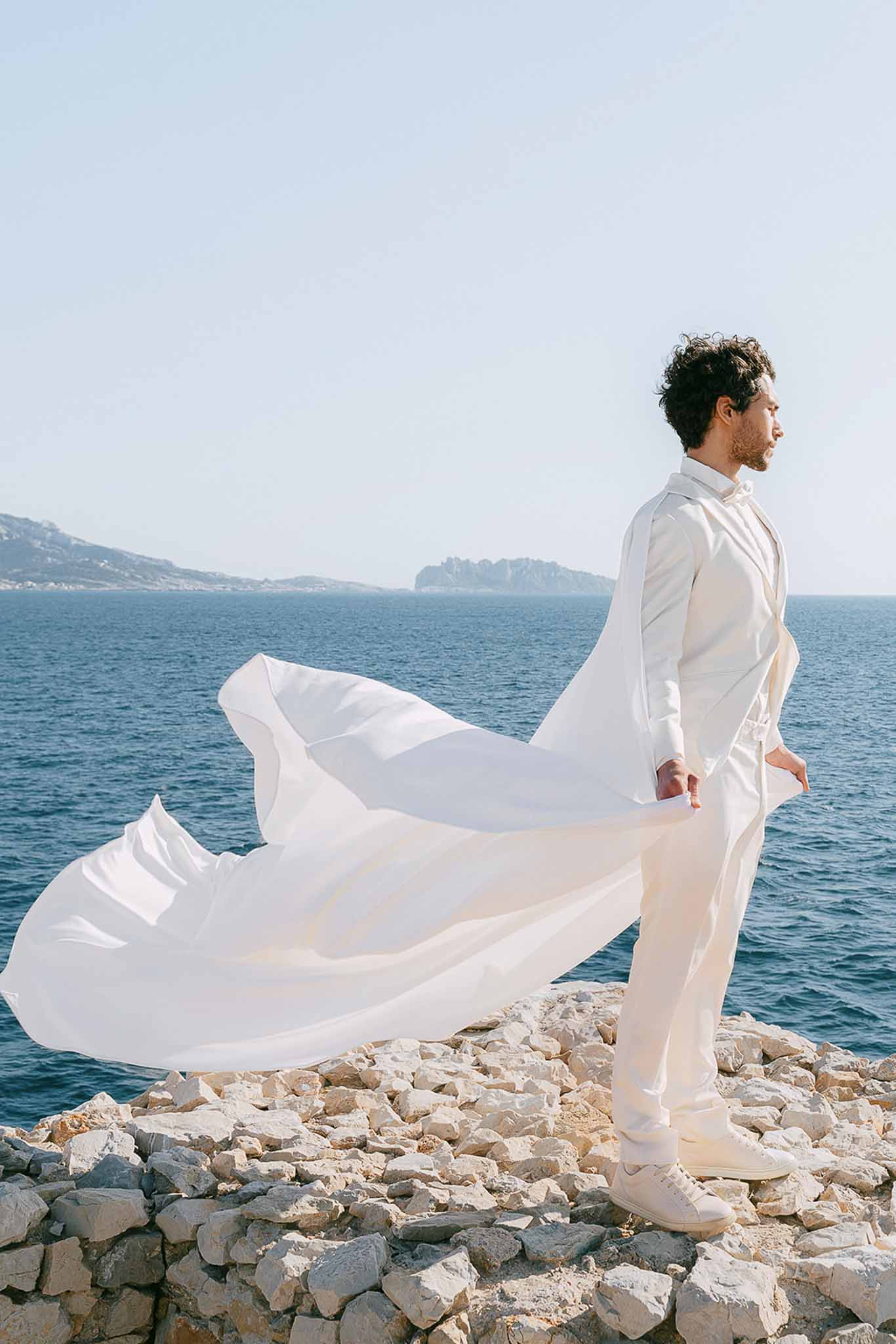 Groom in all-ivory suit with flowing white cape billowing in wind on coastal rocks above blue sea