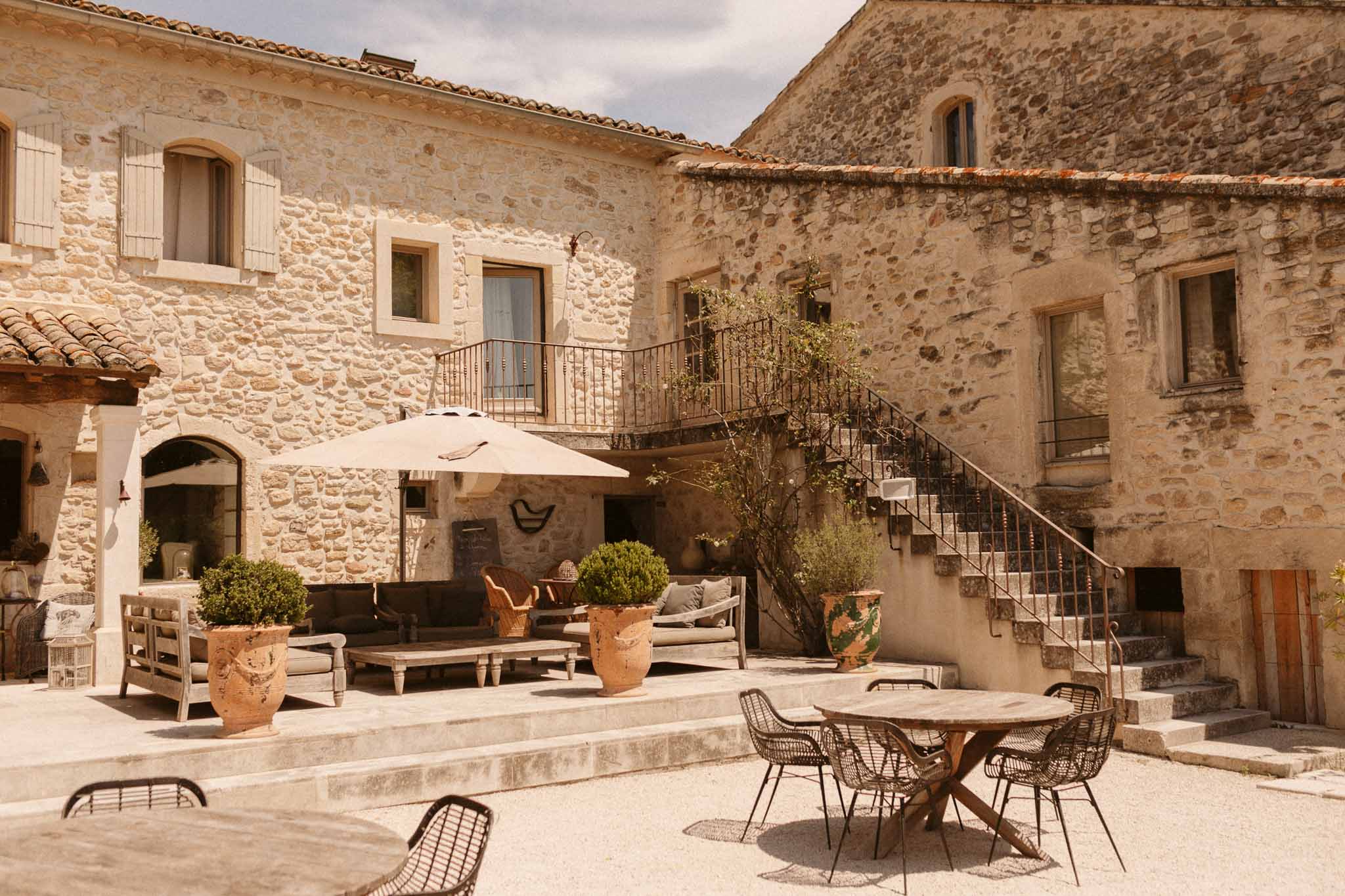Limestone mas courtyard with daybed terrace, topiary urns, wrought-iron staircase, and gravel seating area