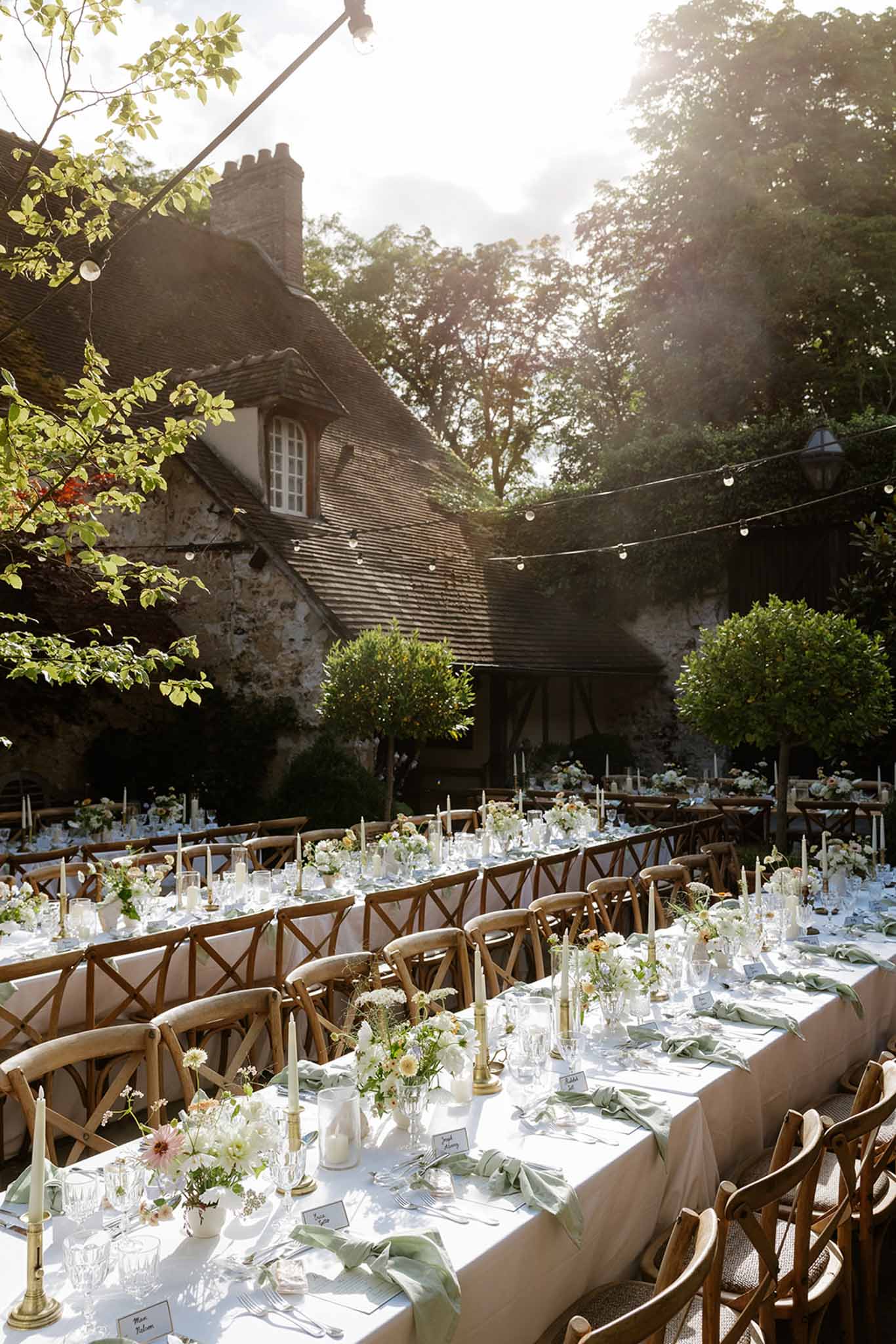 Long tables with sage napkins and peach floral centerpieces under string lights before half-timbered manor courtyard