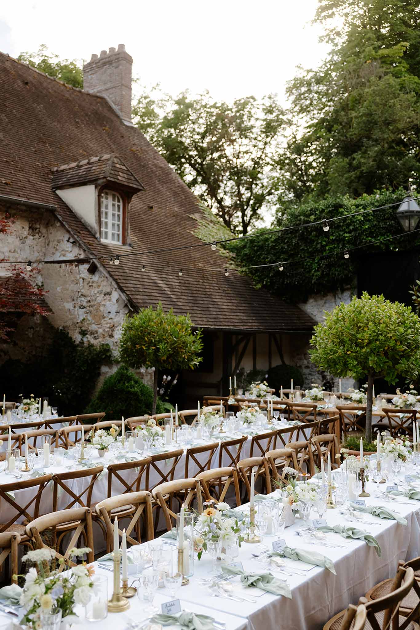Banquet tables with sage napkins, gold tapers, and white floral runners under globe lights at stone manor