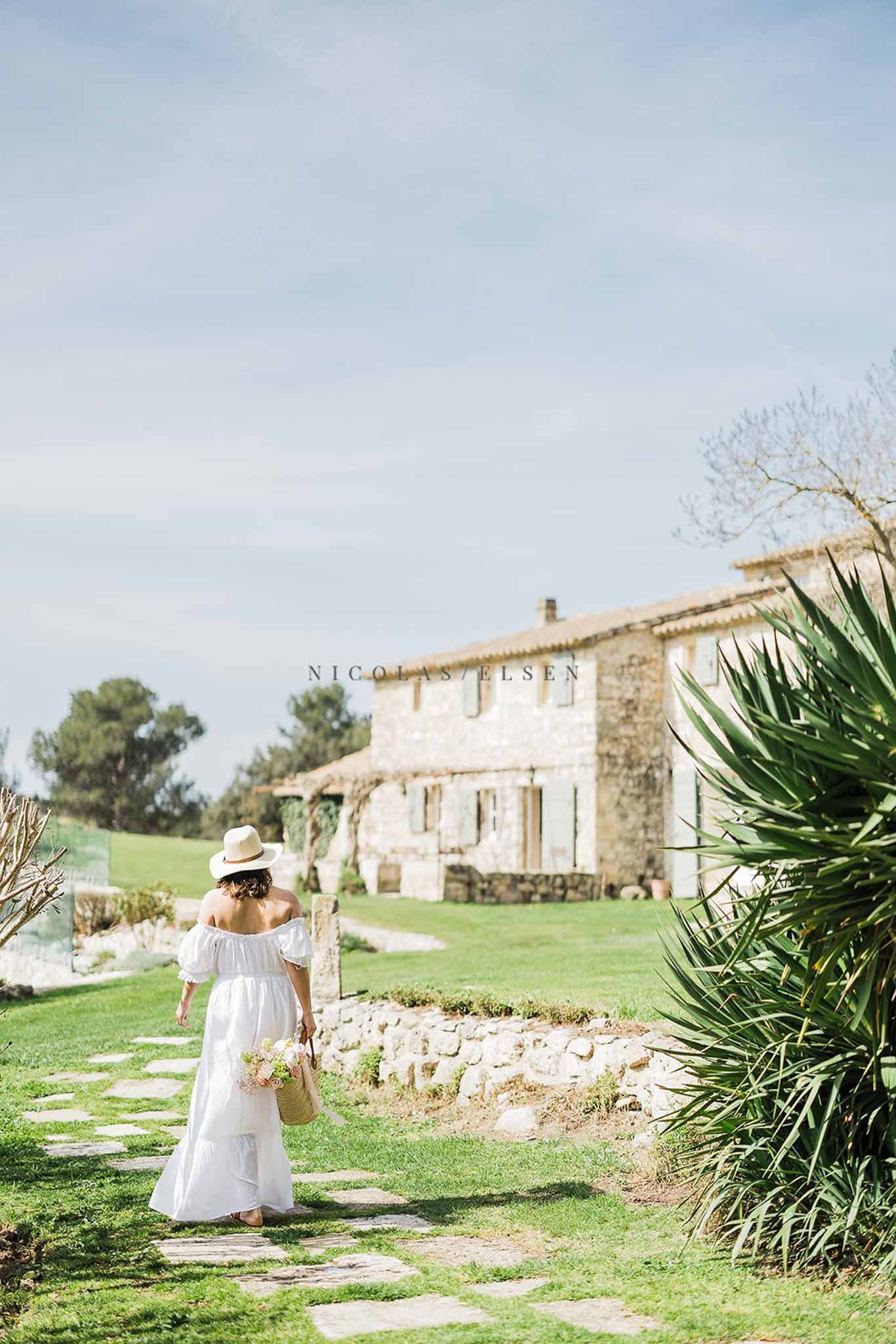 Woman in white off-shoulder dress and straw hat walking along stone path toward Provencal farmhouse