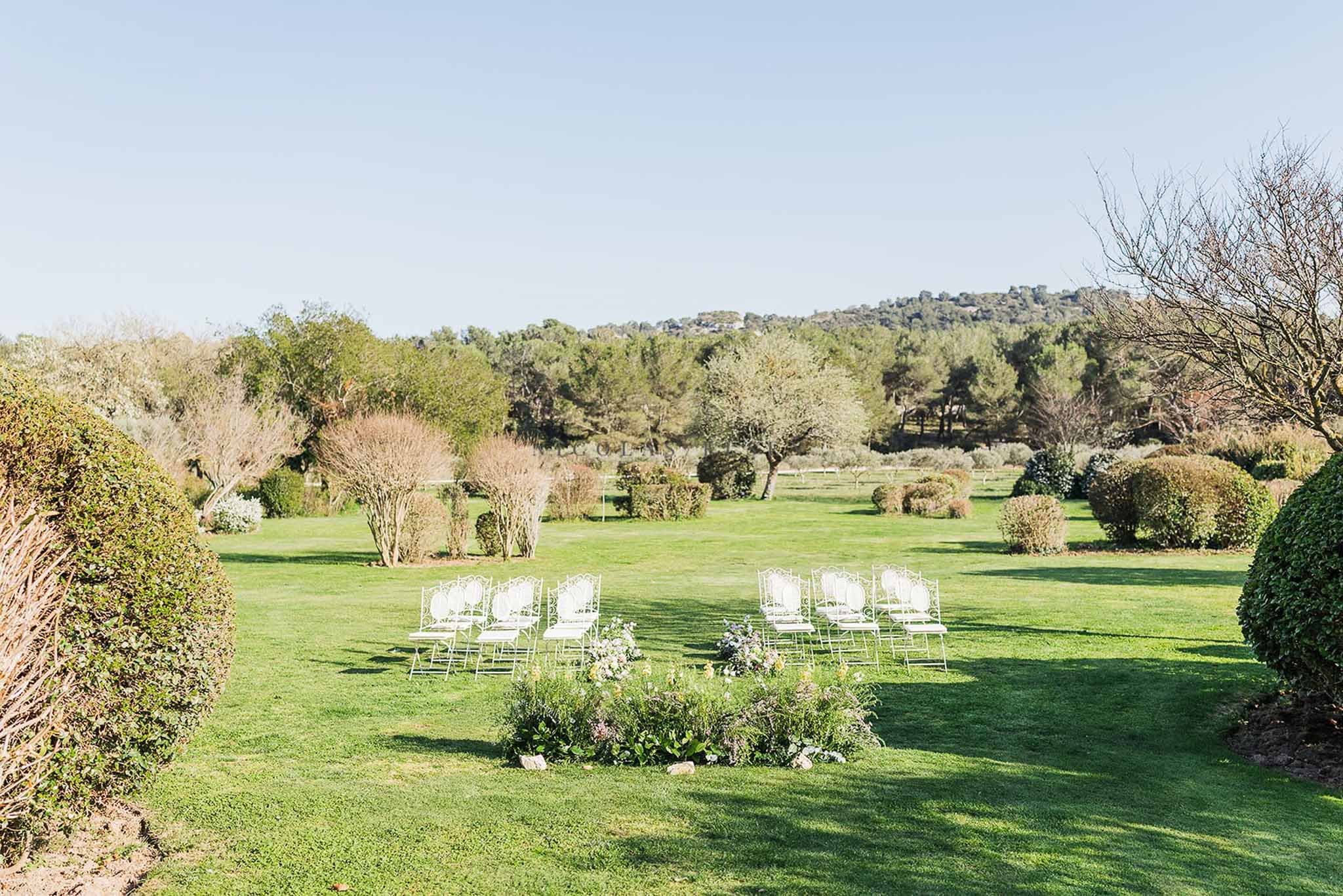 Outdoor ceremony setup with white metal garden chairs in a semicircle and yellow, purple, and white floral ground arrangement