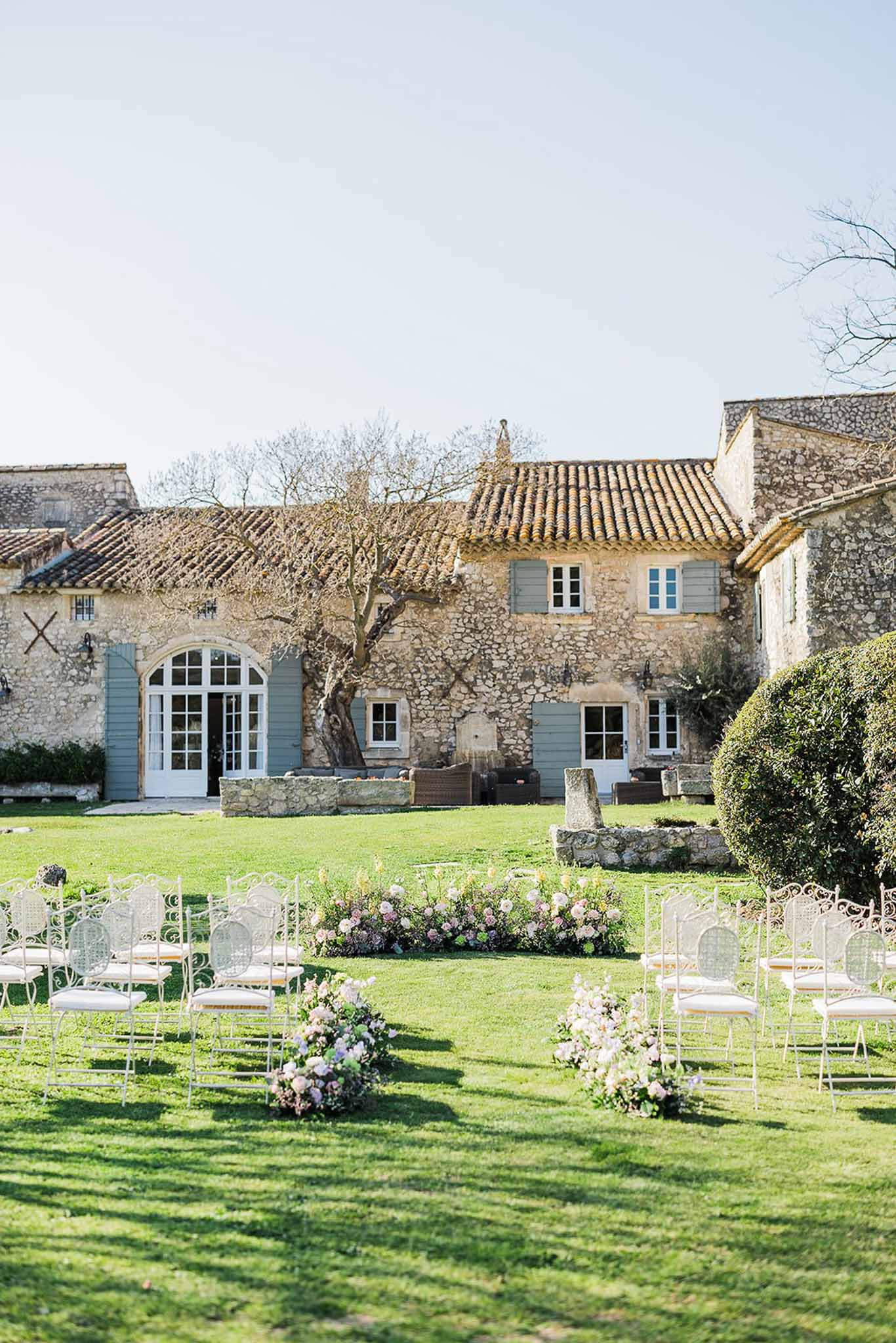 White metal chairs with pastel floral aisle arrangements before Provencal stone farmhouse with blue-grey shutters