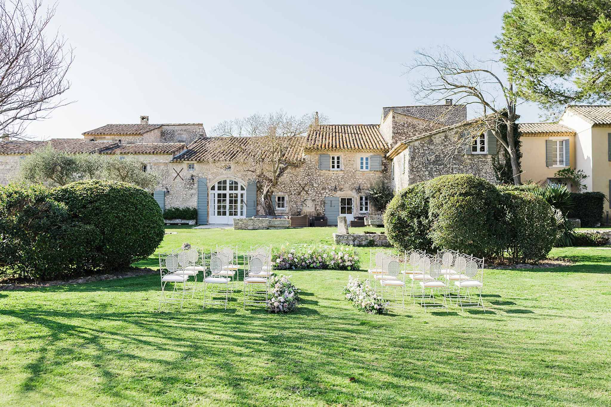 White wrought-iron chairs with pastel floral markers before stone mas with blue-grey shutters on lawn