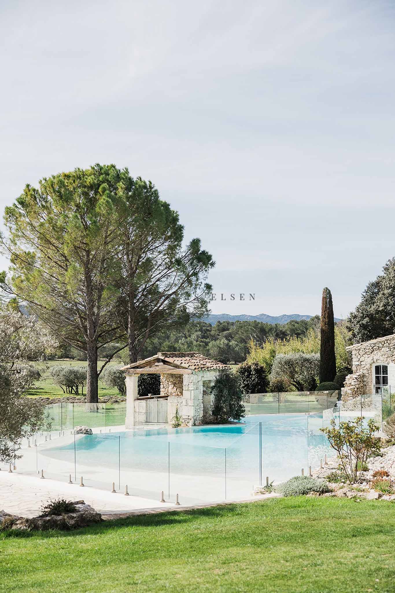 Swimming pool with glass fence, stone cabana, cypress tree, and Provencal outbuildings with mountain backdrop