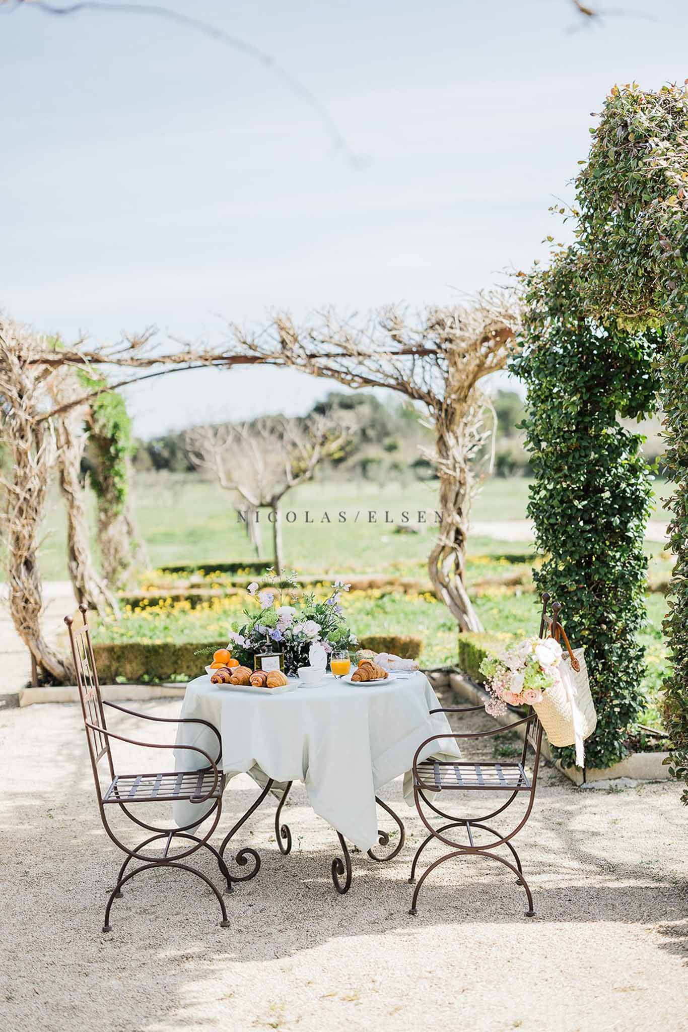 Styled breakfast table with croissants, oranges, and lavender florals under branch pergola in garden