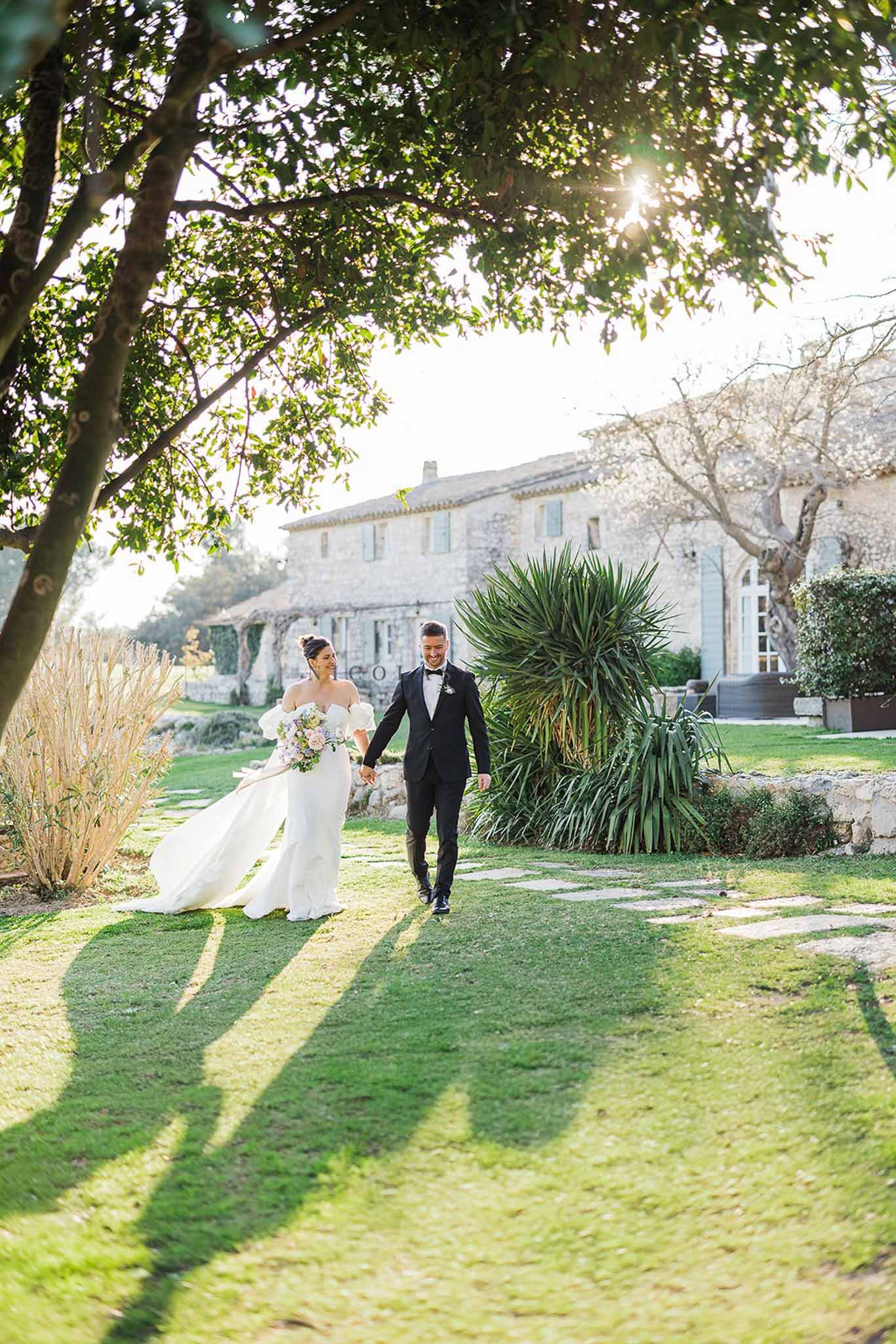 Couple walking hand-in-hand on stepping stones with blush bouquet and stone bastide in warm late-afternoon backlight