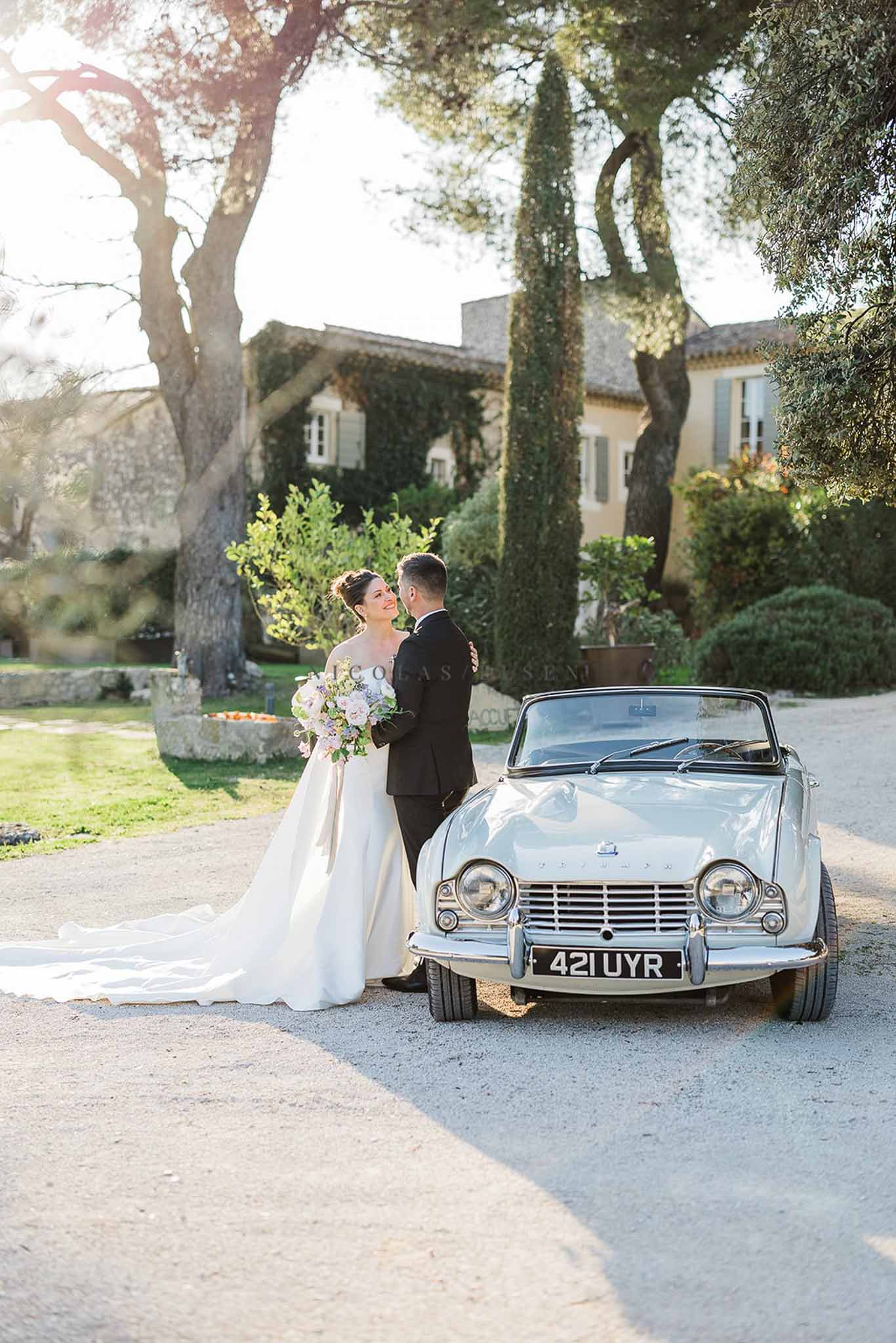 Bride and groom with blush bouquet beside white Triumph convertible at ivy-covered Provencal manor