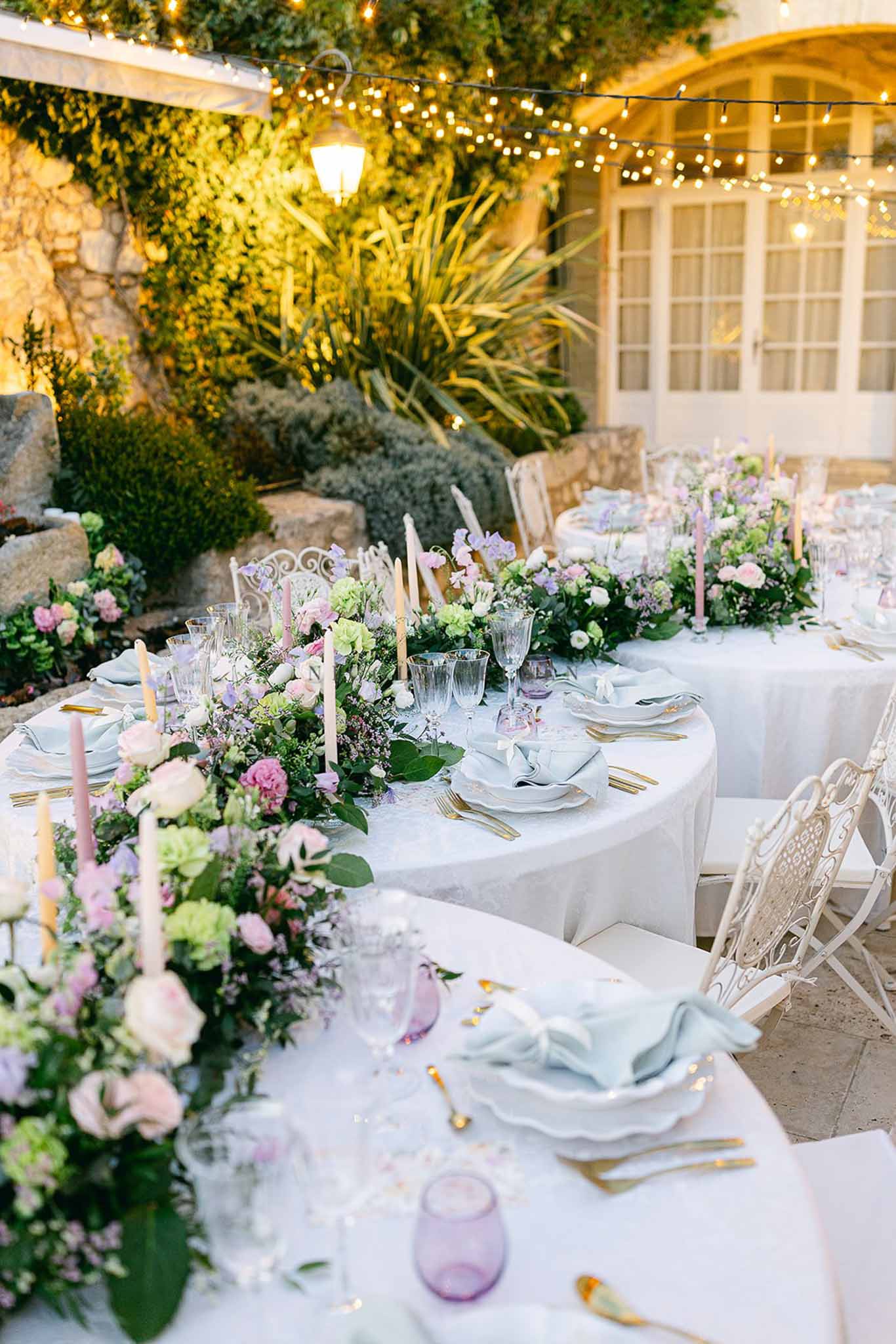 Long tables with pastel floral garland of blush roses and lilac sweet peas under fairy lights in courtyard