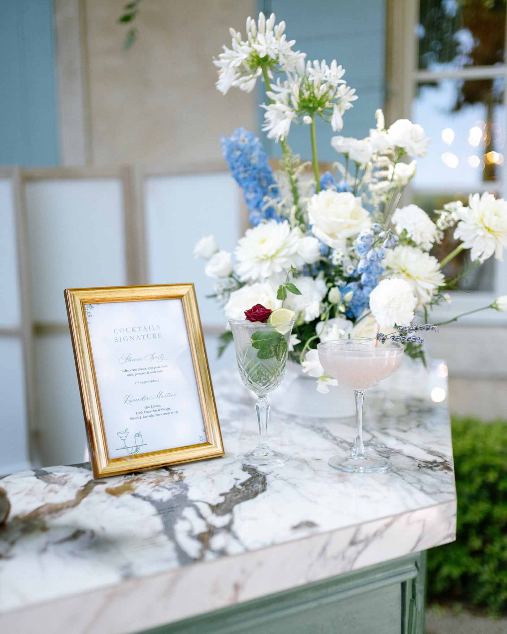 Sage green cocktail bar with signature drink menu, crystal glasses, and blue delphinium floral arrangement