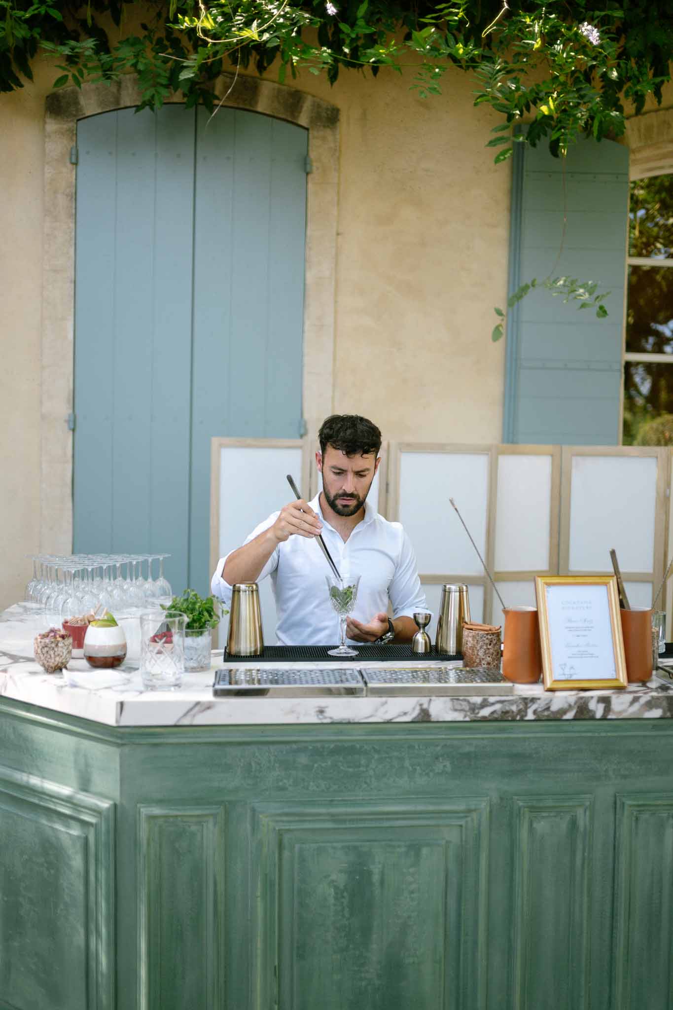 Bartender prepares cocktail at sage green bar with marble top during outdoor wedding cocktail hour at Provencal venue