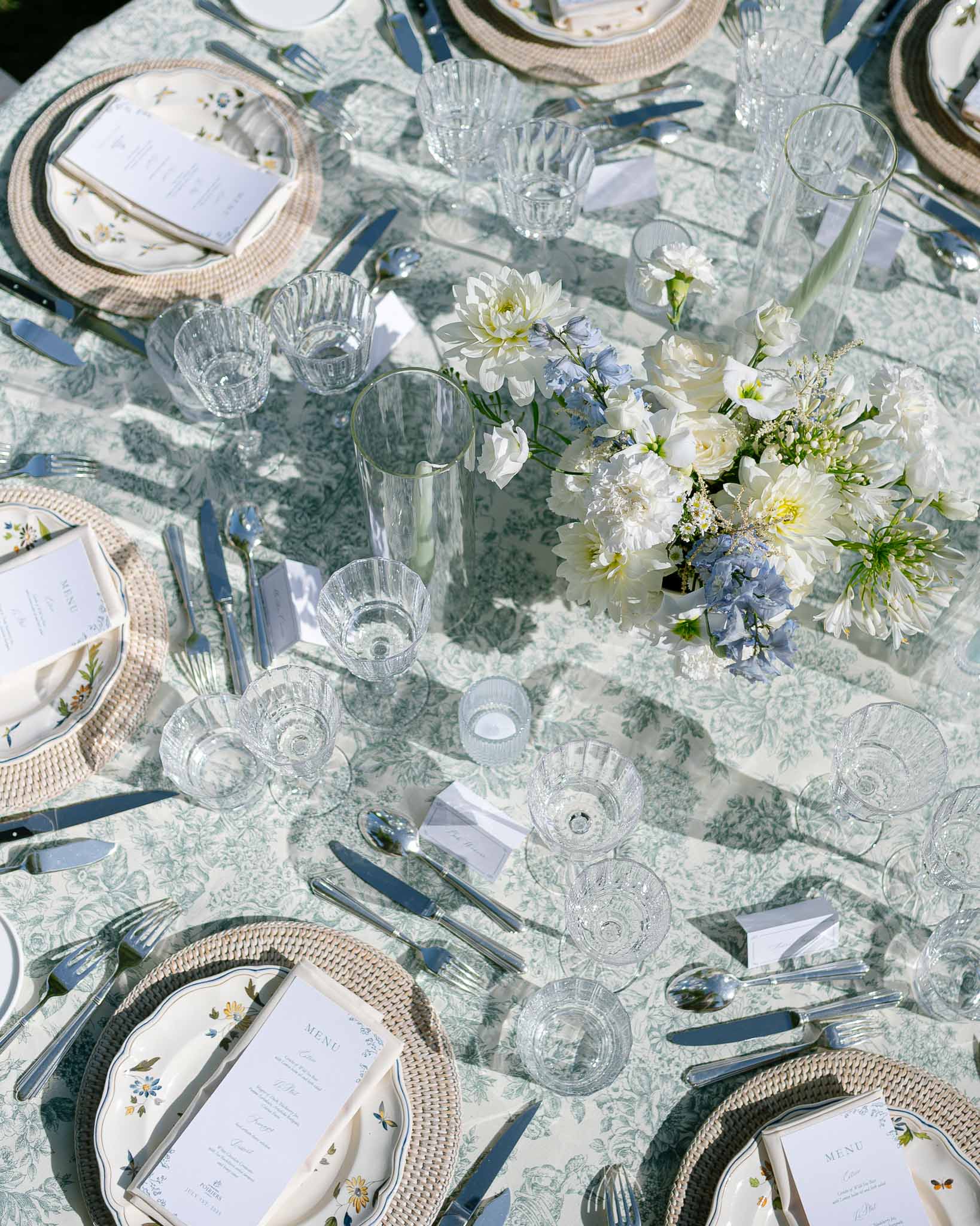 Overhead view of reception table with sage green toile de Jouy tablecloth, floral porcelain plates, and white dahlia cente...