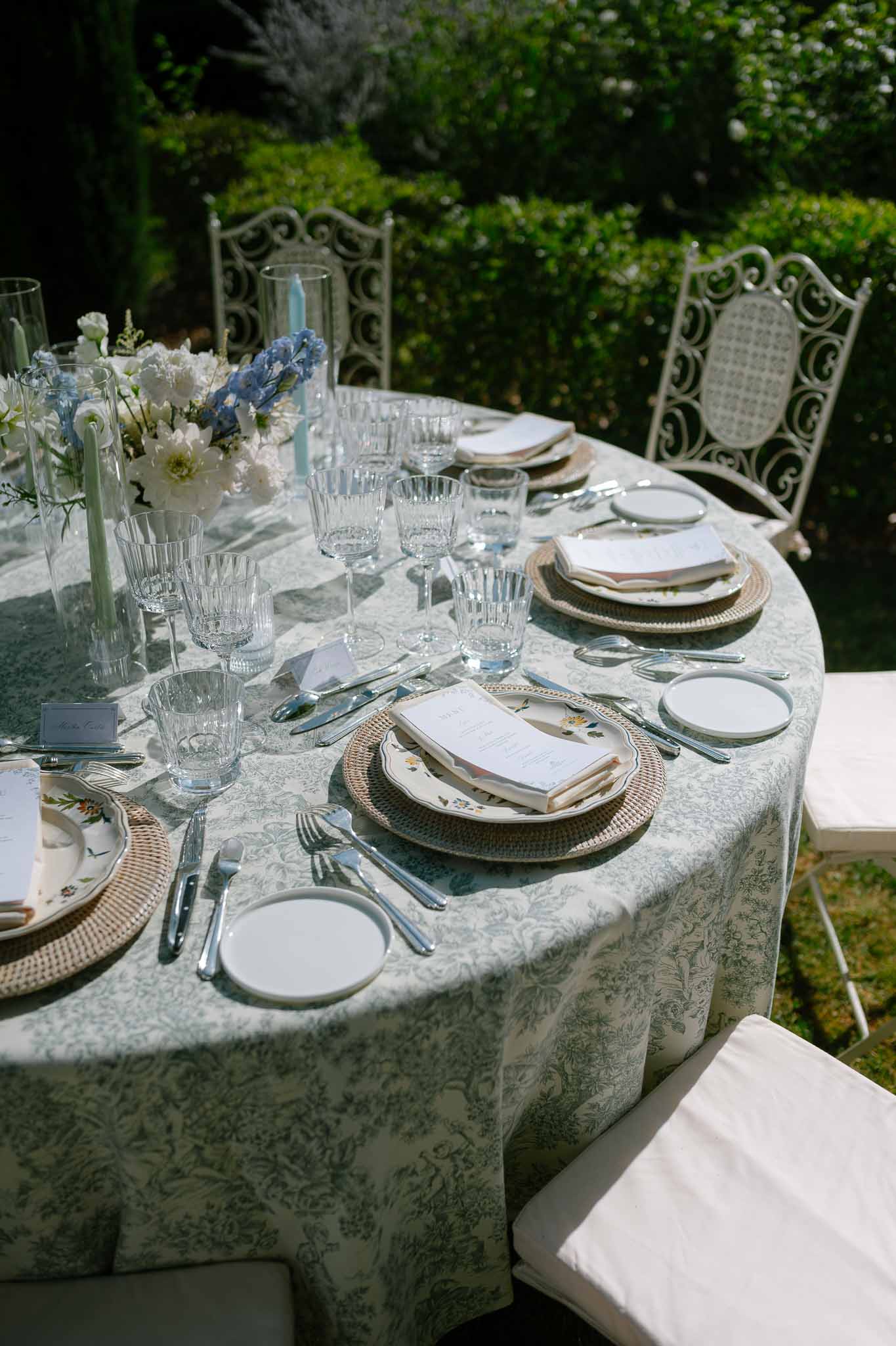 Garden reception table with sage green toile tablecloth, rattan chargers, white dahlias and blue delphiniums centerpiece