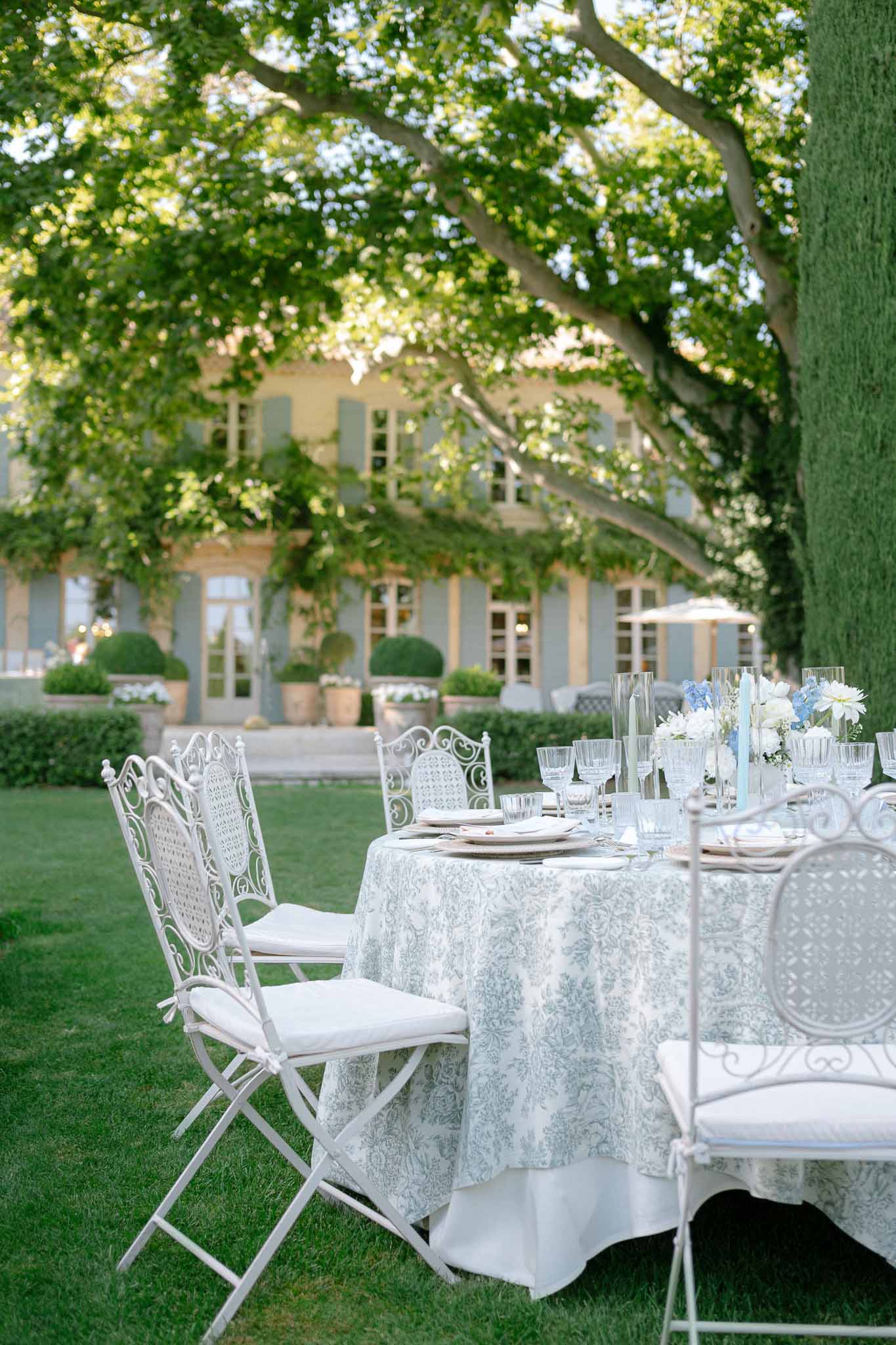 Round table with blue toile tablecloth, white dahlias, and iron chairs before Provencal manor