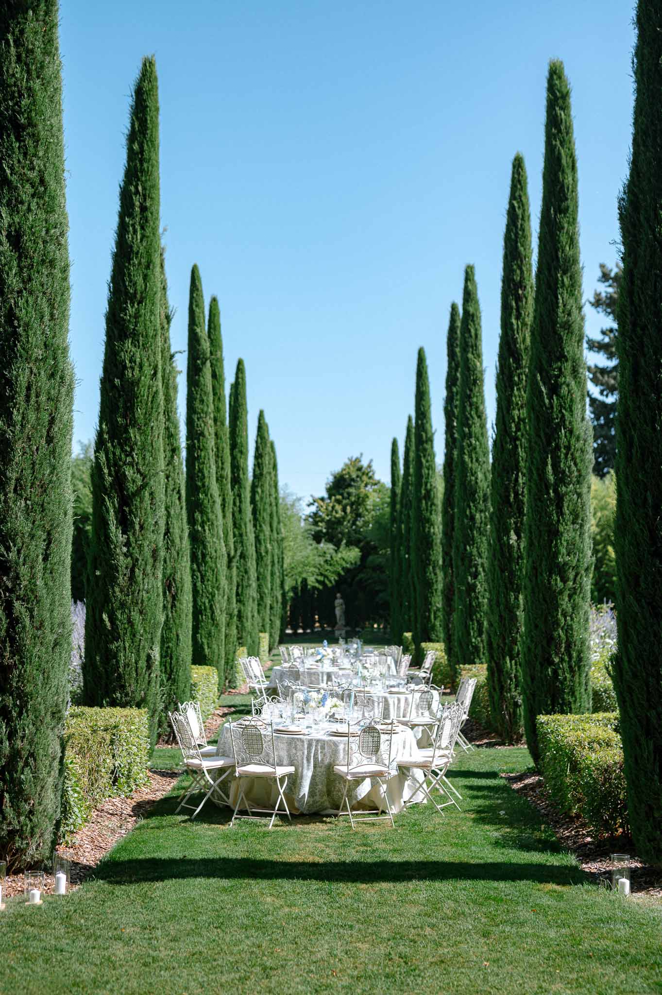 Outdoor wedding reception tables lined along cypress tree alley with white wrought-iron chairs and floral centerpieces