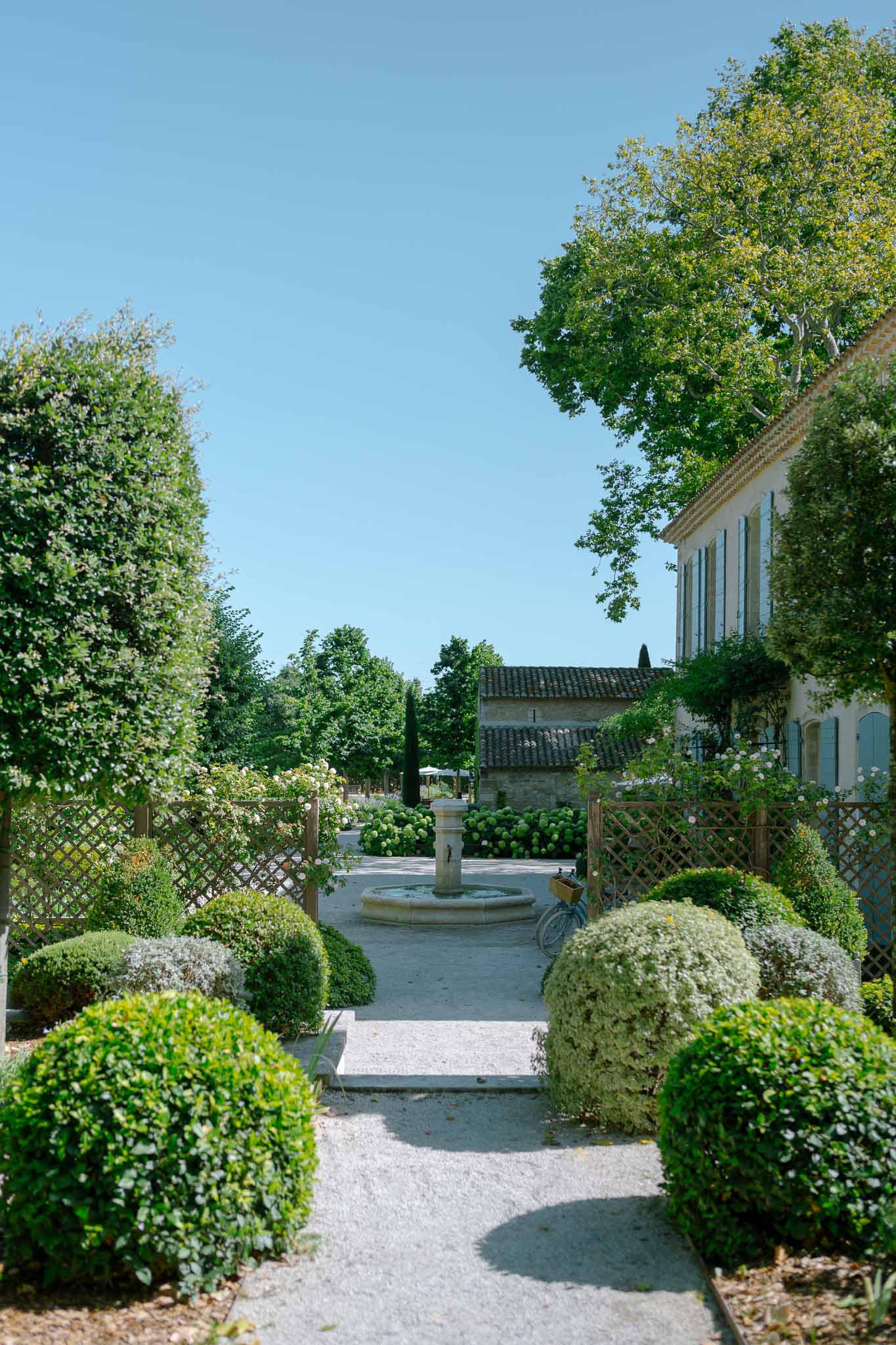 Provencal garden with box ball topiaries, stone pedestal fountain, and two-storey mas with blue shutters