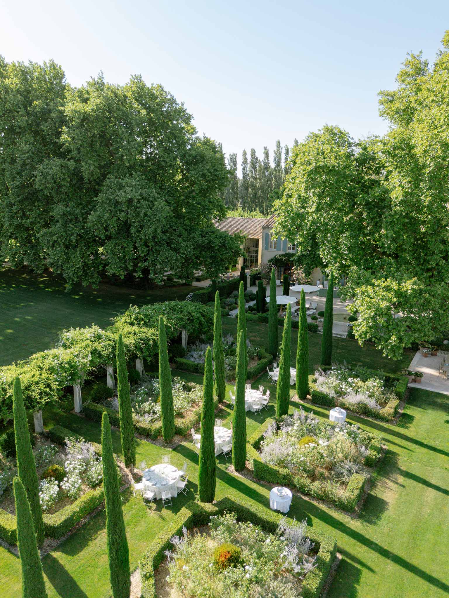 Aerial garden reception with round tables between cypress trees white folding chairs and Provencal manor behind