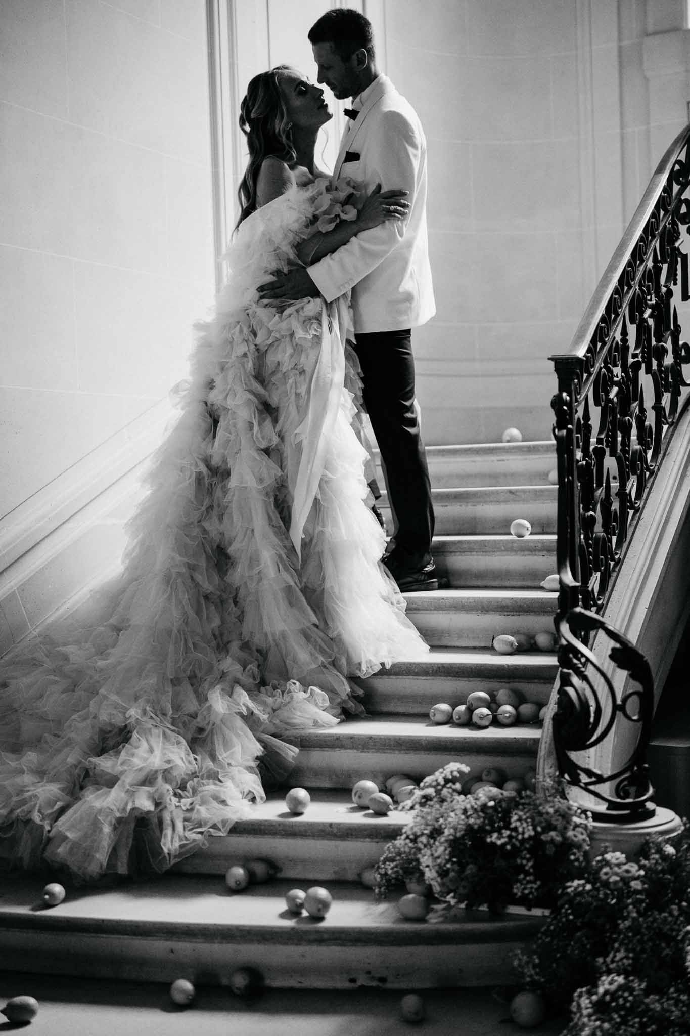 Black and white portrait of bride and groom embracing on grand chateau staircase with ruffled tulle train cascading down s...