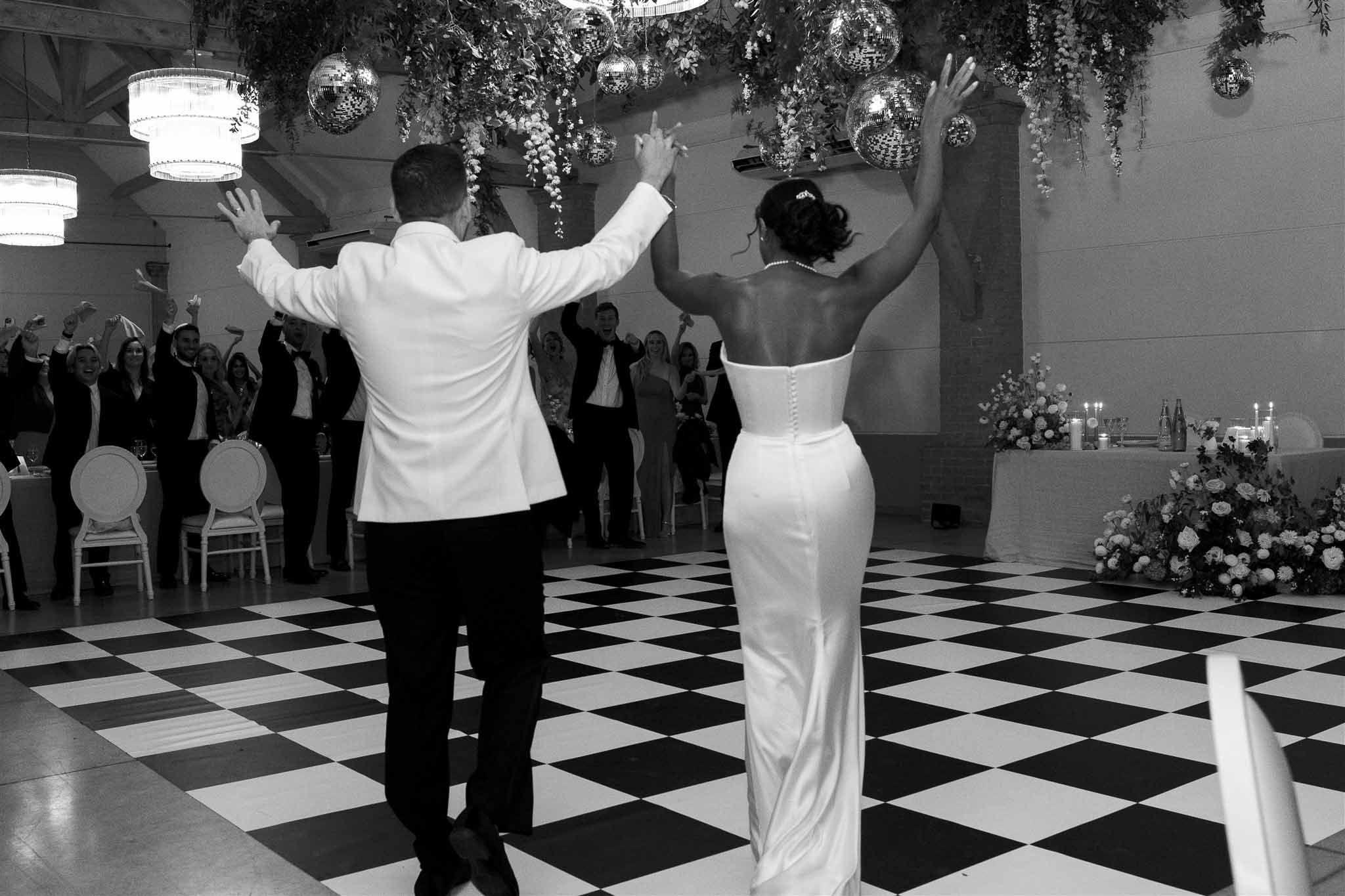 Black and white shot of bride and groom with arms raised on checkered dance floor with hanging greenery and disco balls