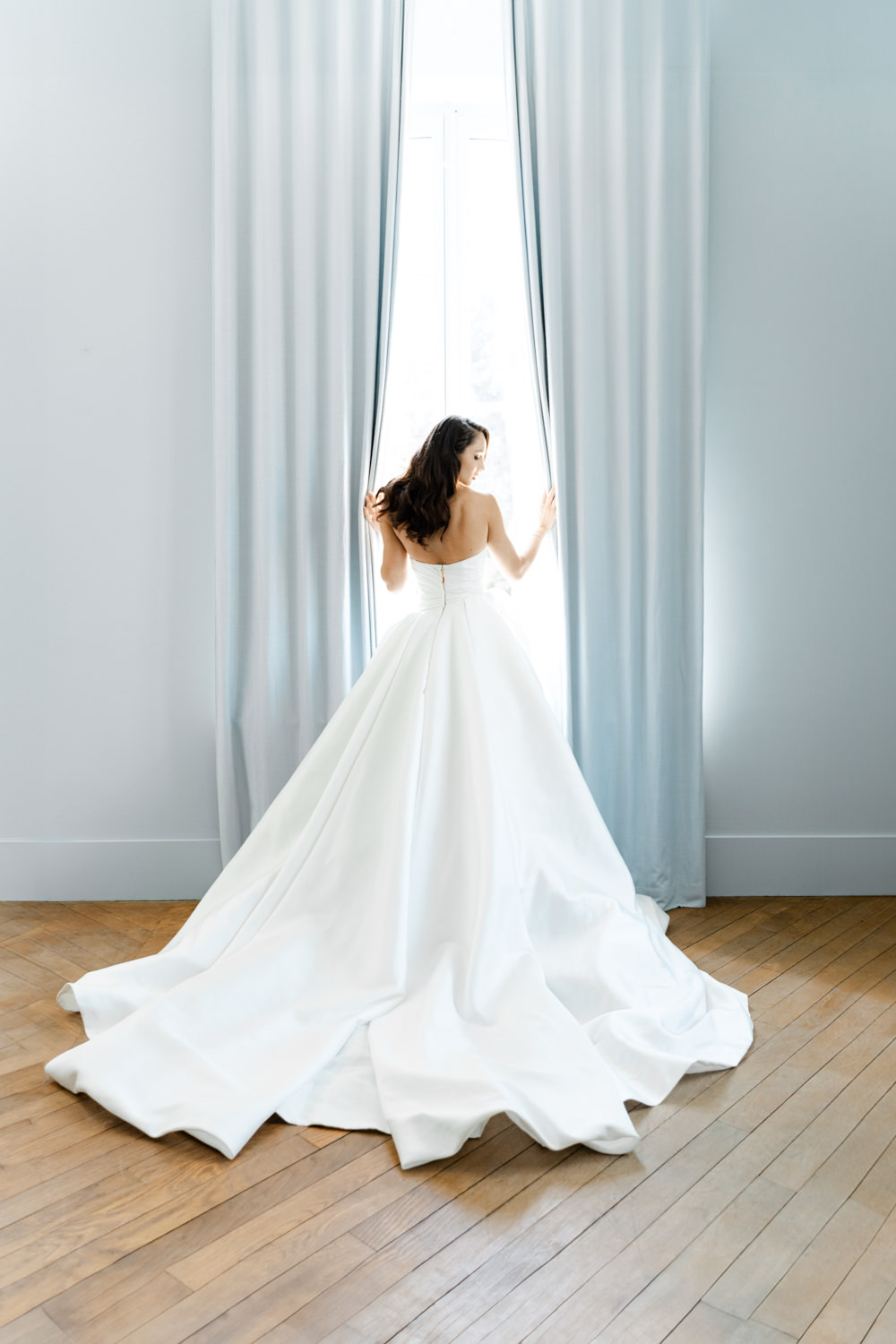 Bride from behind holding blue-grey curtains open with cathedral train spread across parquet floor