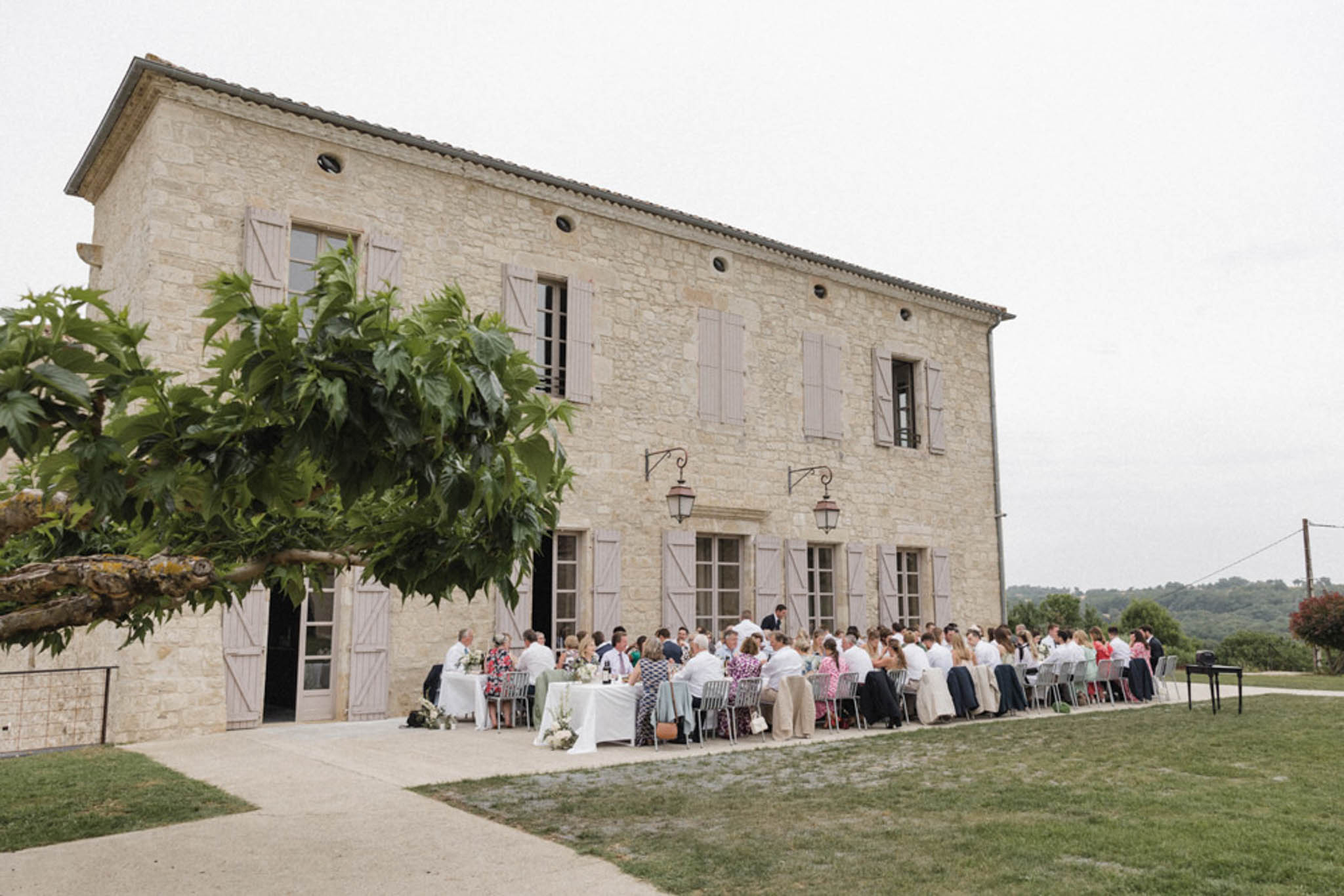 Fifty guests at long table before Provencal stone farmhouse with taupe shutters and wrought-iron lanterns