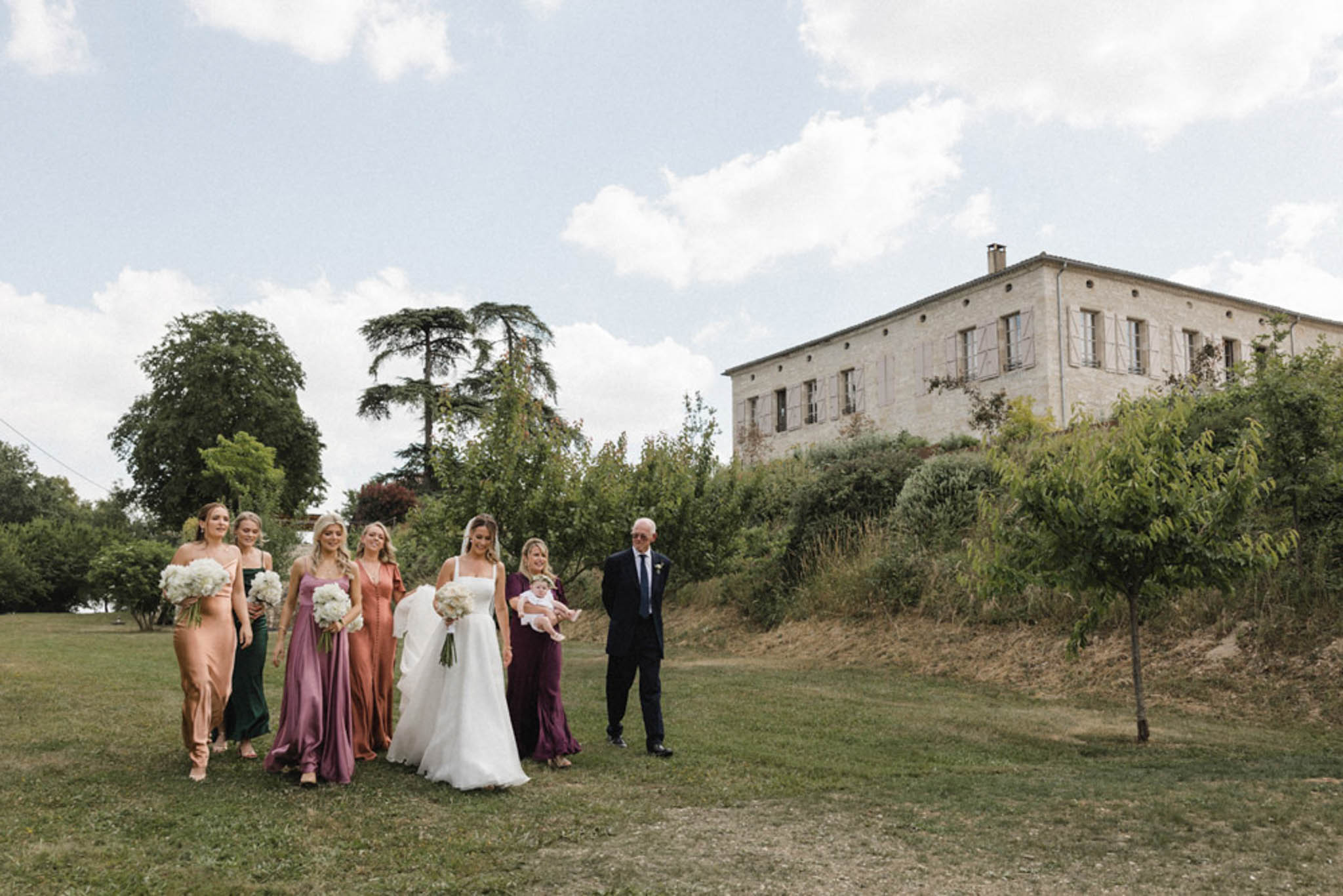 Bride and five bridesmaids in mismatched green terracotta mauve and plum dresses walking across manor lawn