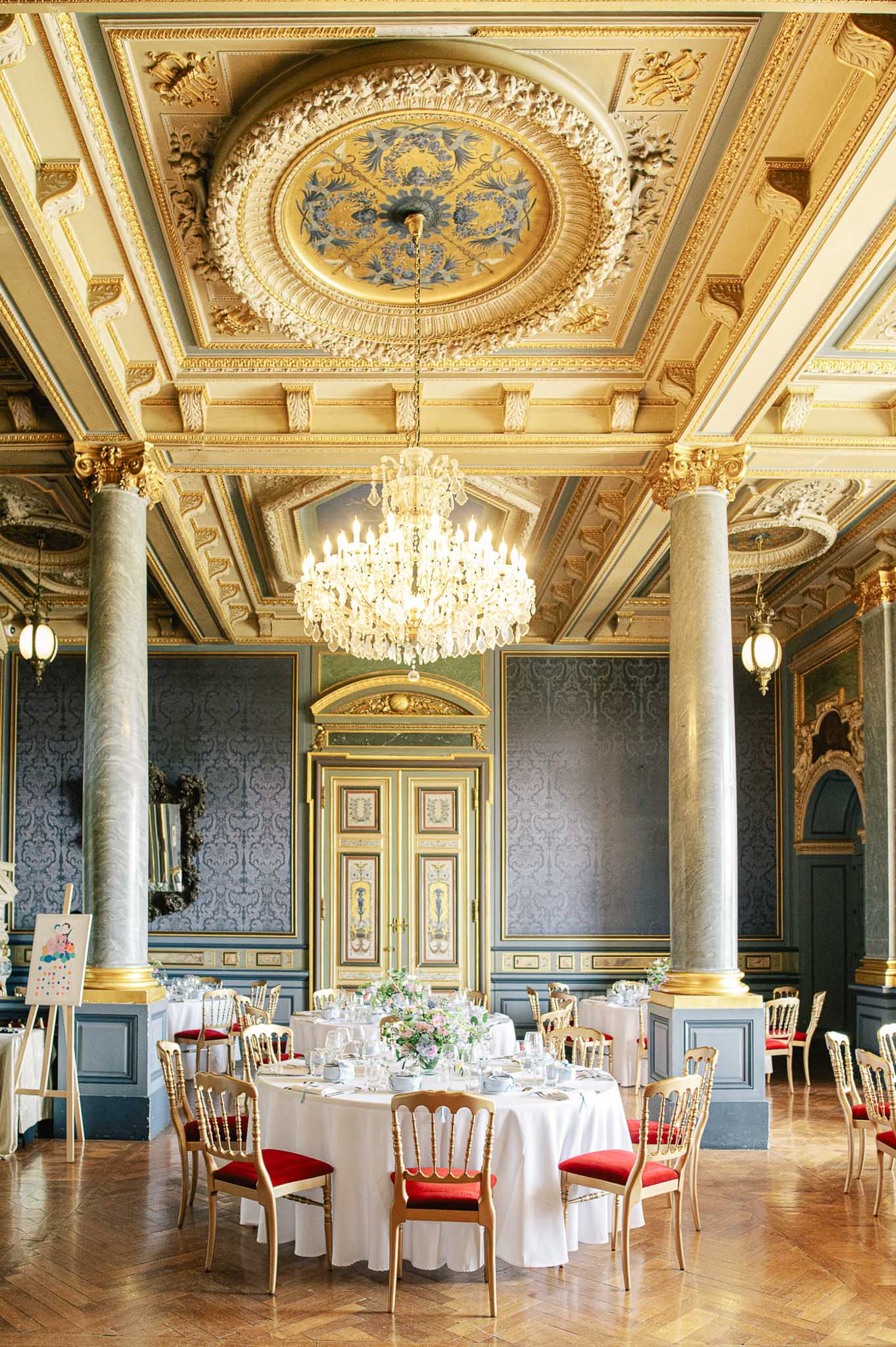 Grand ballroom with gilded ceiling, crystal chandelier, and round tables set with pink florals