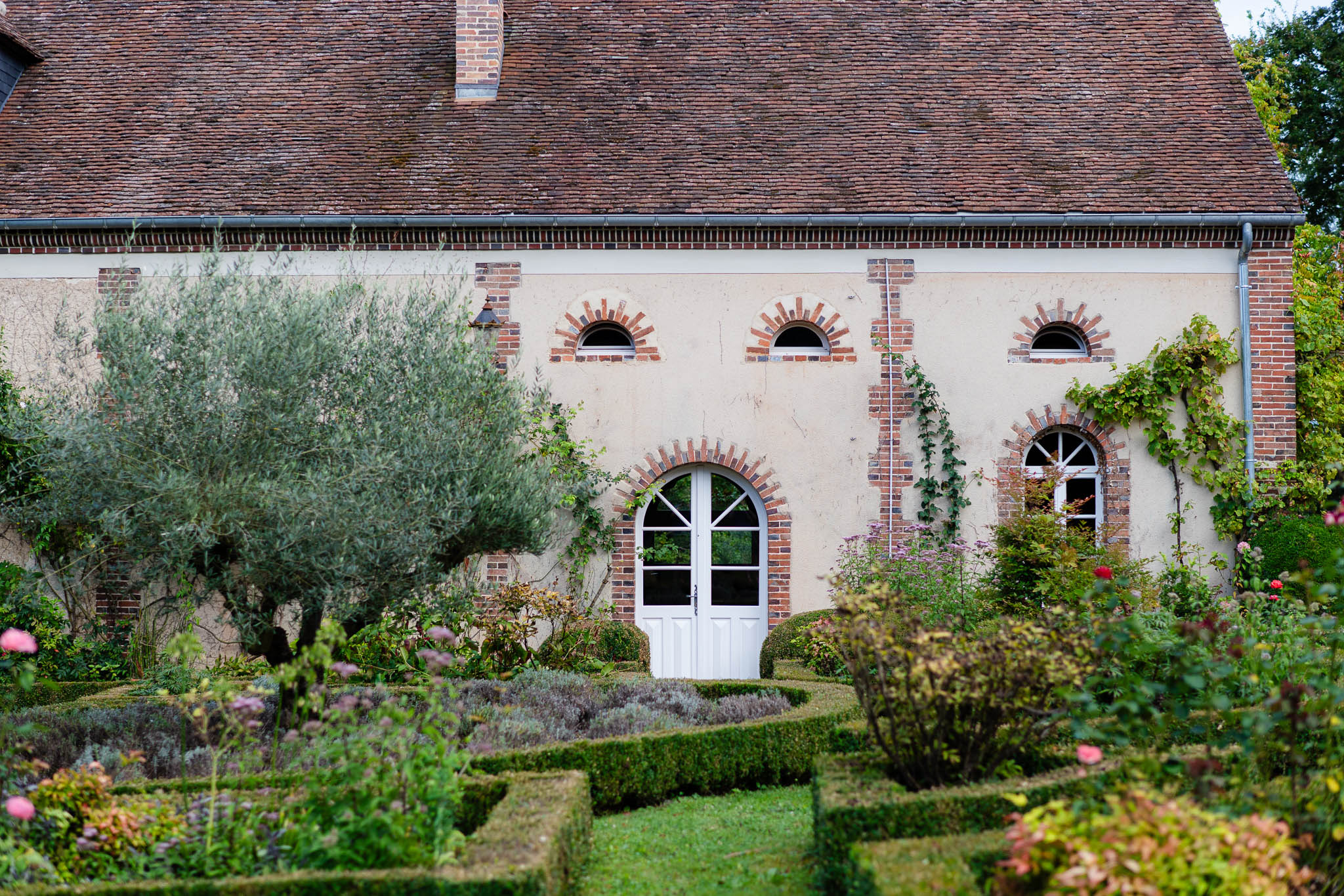 French country manor facade with cream walls, red brick arches, and formal parterre garden with boxwood hedges
