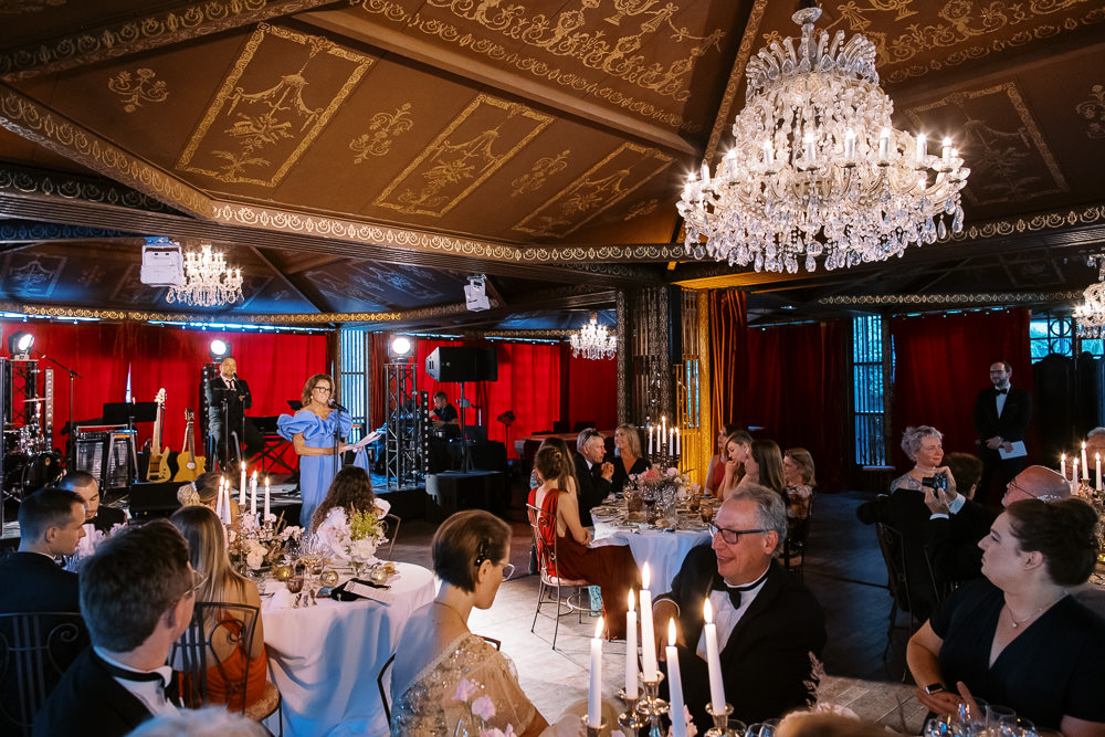 Wedding speech in grand ballroom with wood-paneled ceiling, crystal chandeliers, and round candlelit tables