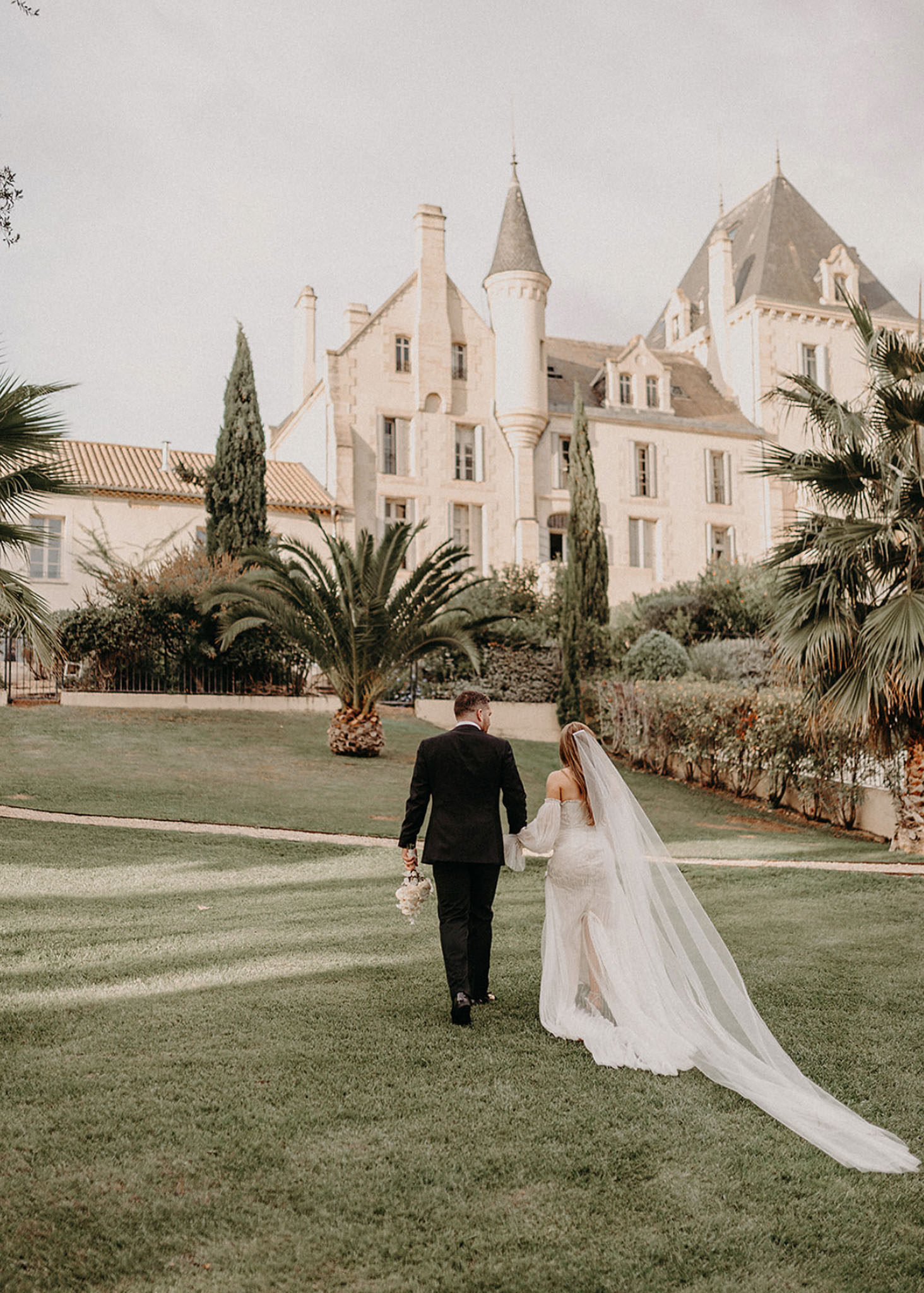 Couple walking toward turreted chateau from behind bride in off-shoulder lace gown with cathedral veil