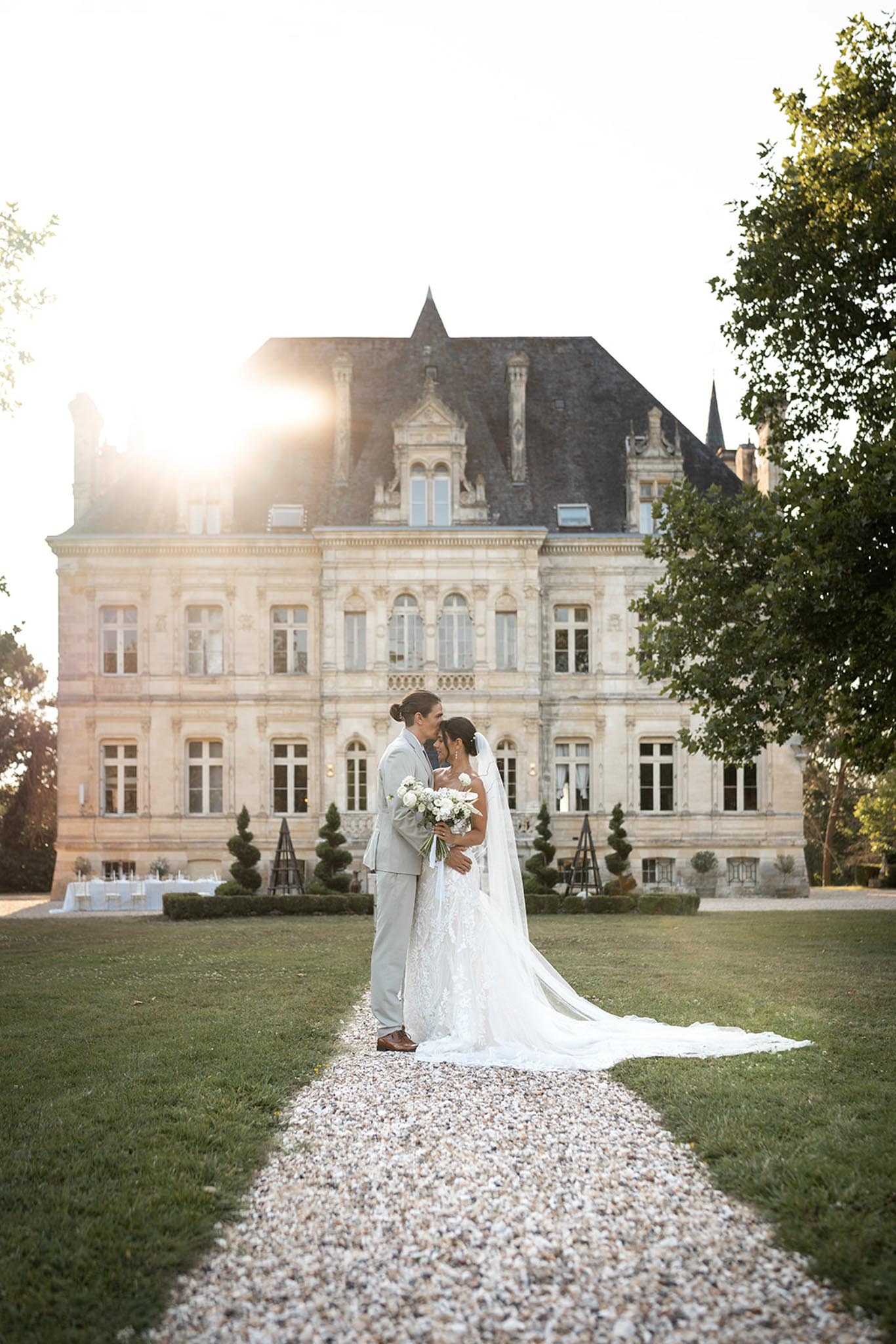 Couple nearly kisses on gravel path toward chateau at golden hour with white rose bouquet and long veil