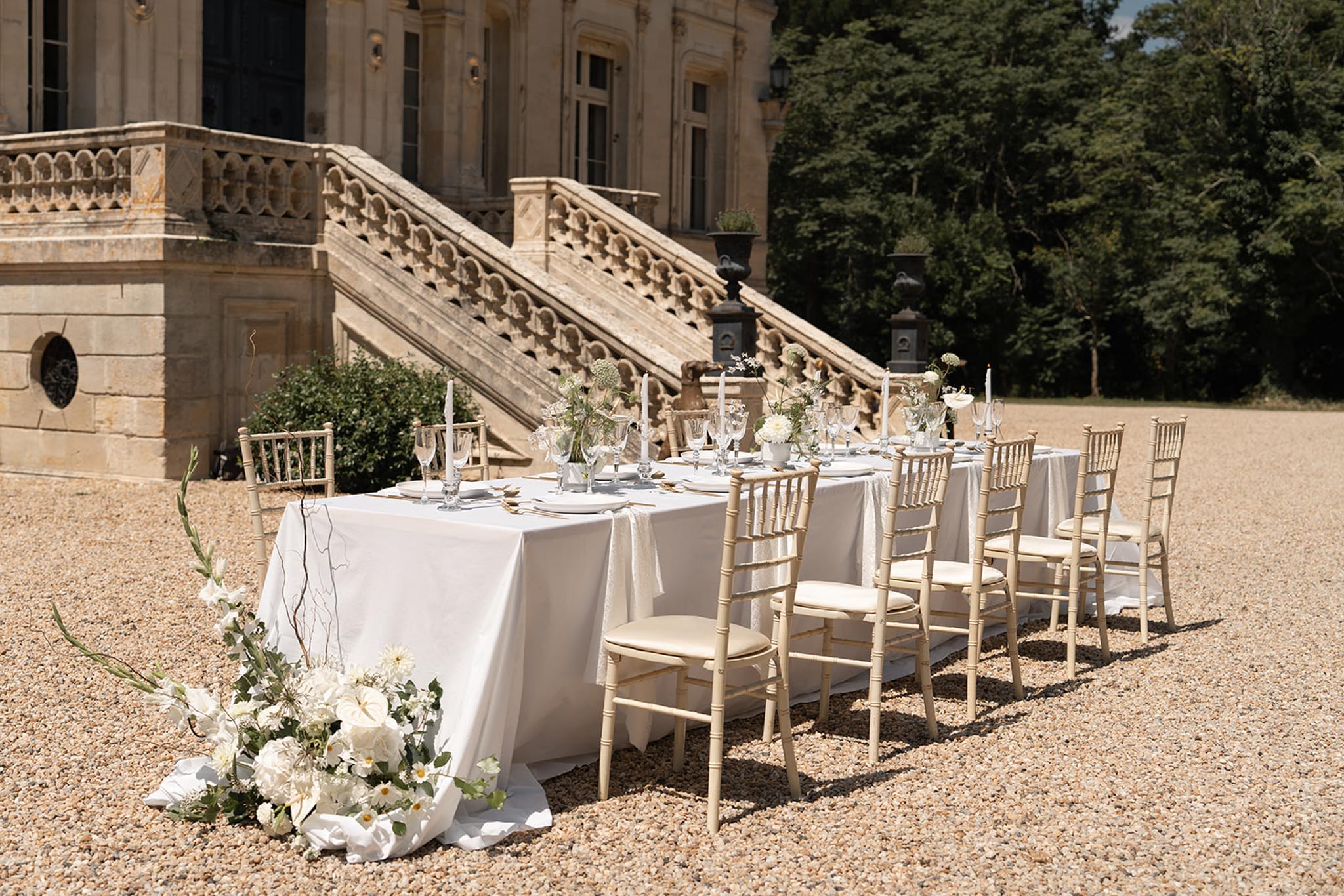 Long reception table with white dahlias and taper candles before chateau stone staircase and urns