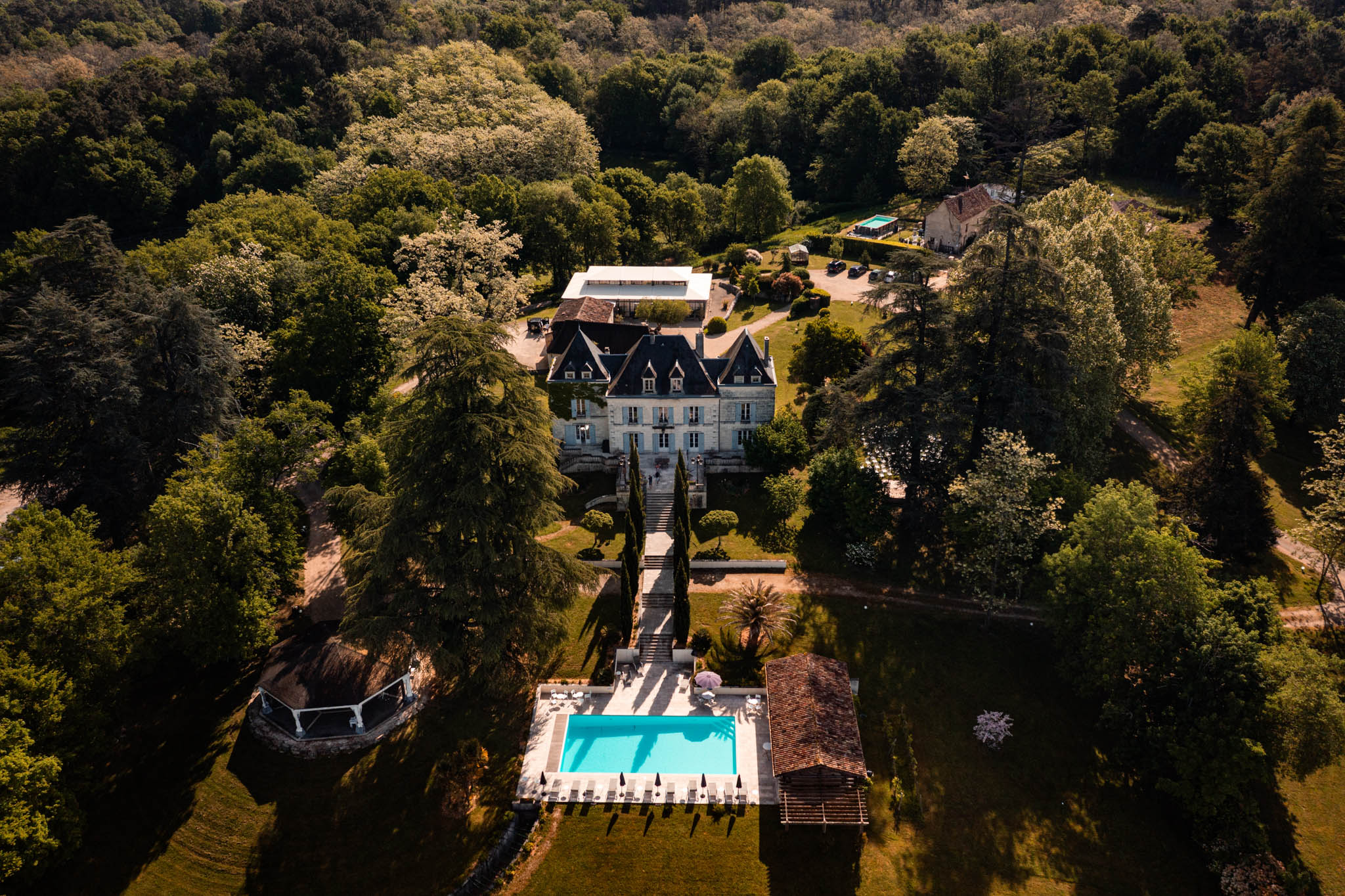 Aerial drone view of French chateau estate with mansard roofs, cypress-lined staircase, and turquoise swimming pool