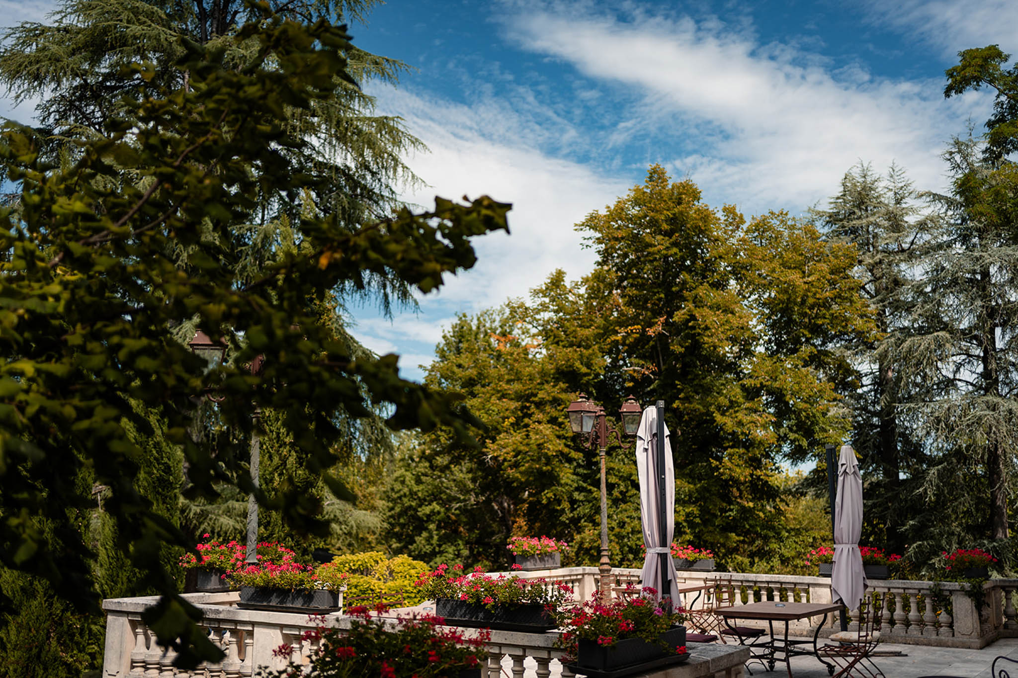 Stone terrace with balustrade, red flowering planters, iron lamp posts, and bistro seating at a chateau