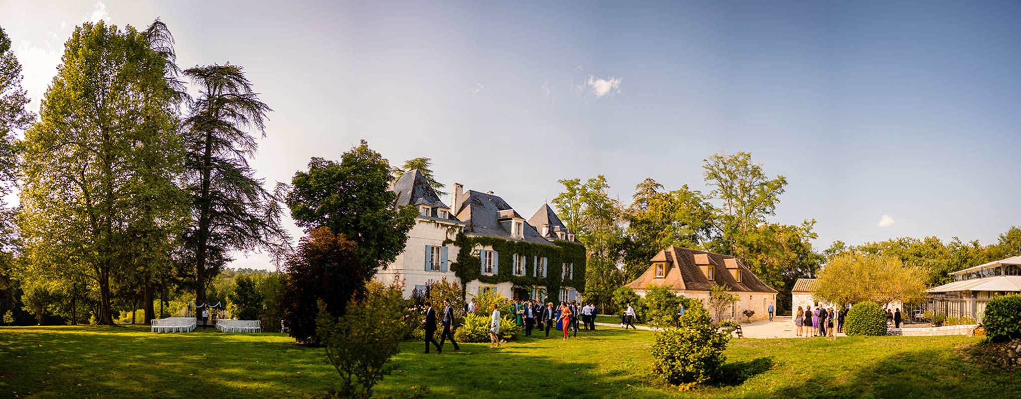 Panoramic view of a white stone chateau with guests gathered on the grounds during golden hour cocktail reception