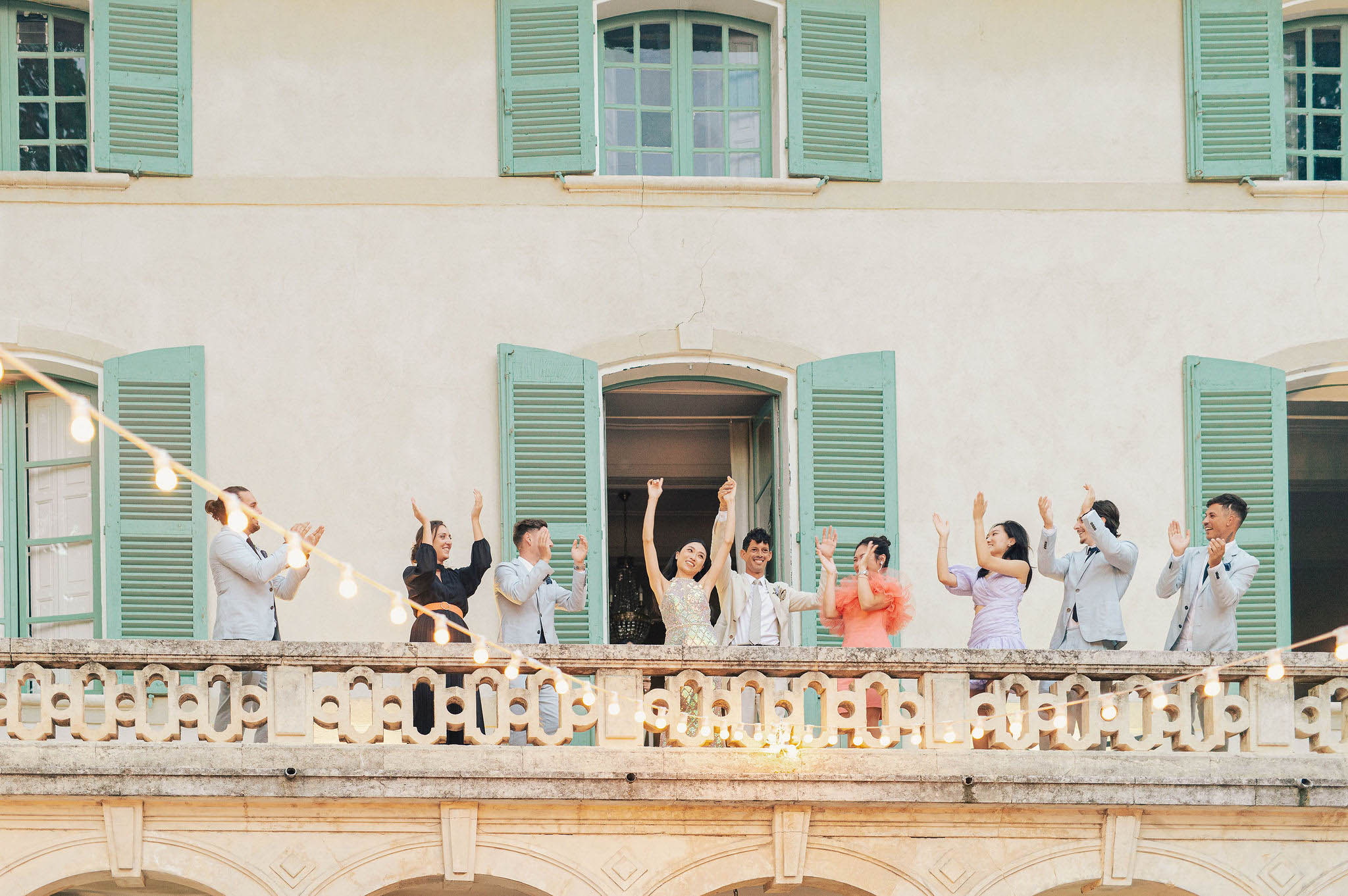 Bride in sequined dress and groom celebrate with guests on chateau balcony with string lights at night