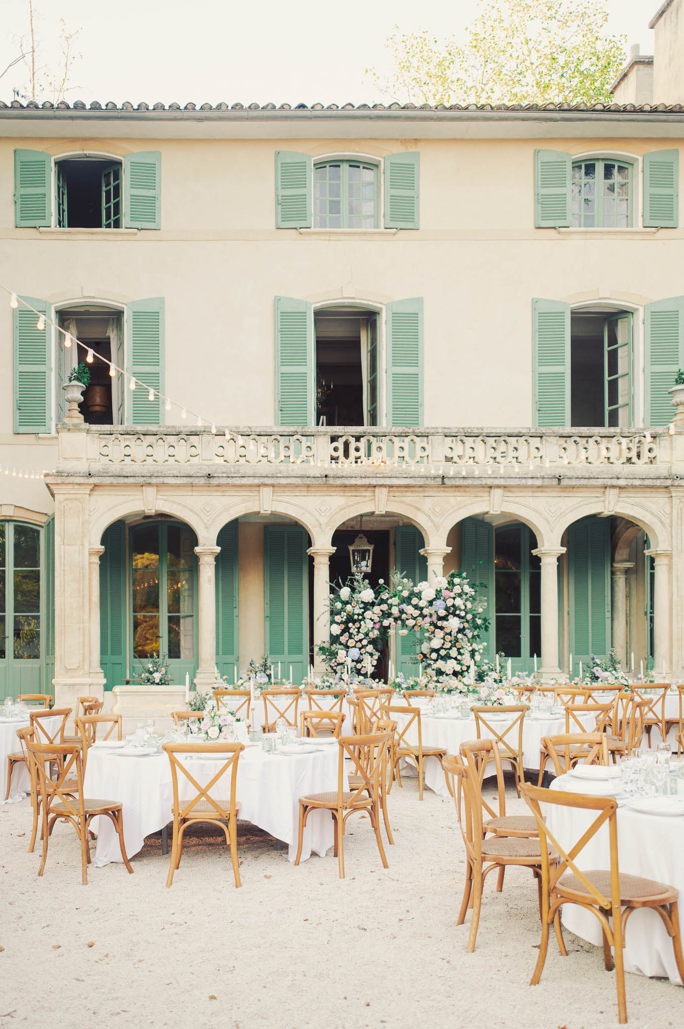 Outdoor courtyard reception with round tables, cross-back chairs, pastel floral centerpieces, and French manor facade with...